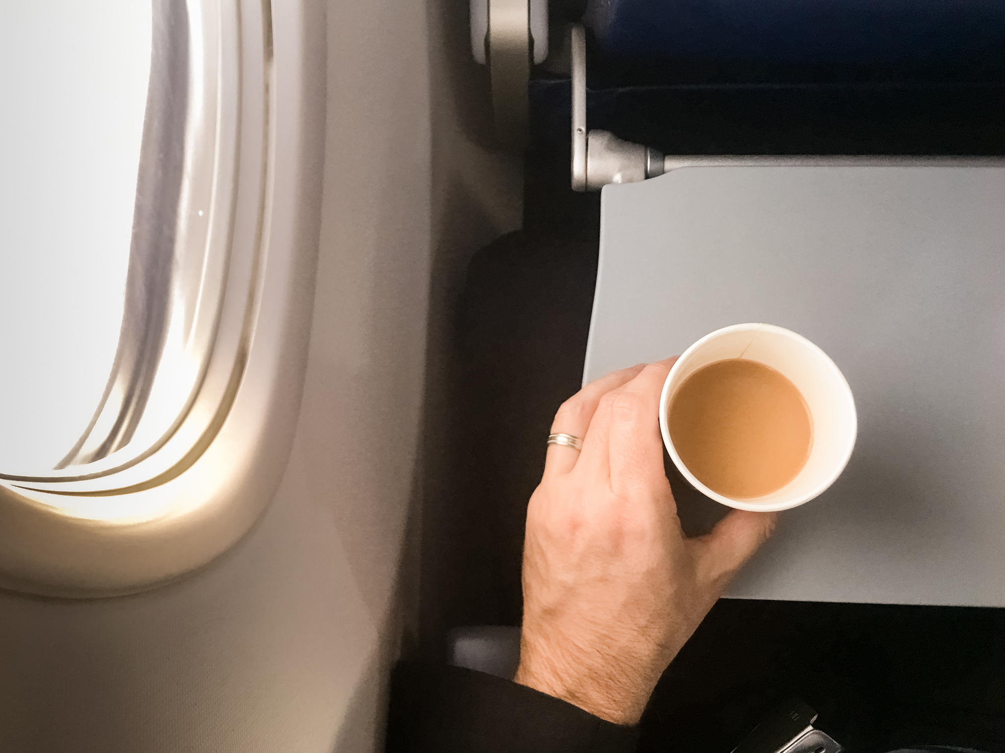 A man's hand with a wedding ring holds a cup of coffee next to an airplane window and seat tray