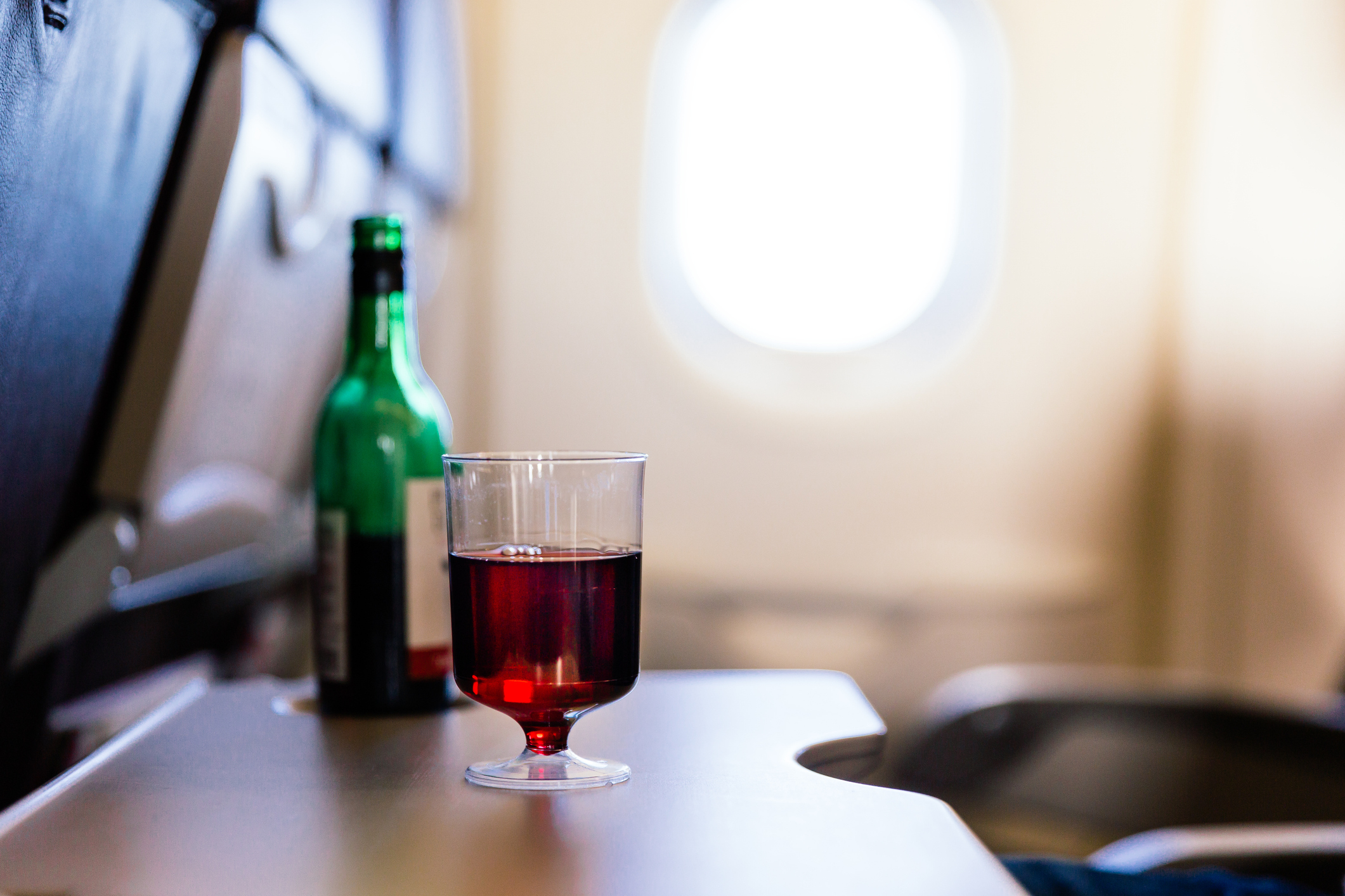 A glass of red wine and a green wine bottle are on an airplane tray table near a window. The background shows an airplane seat and a window