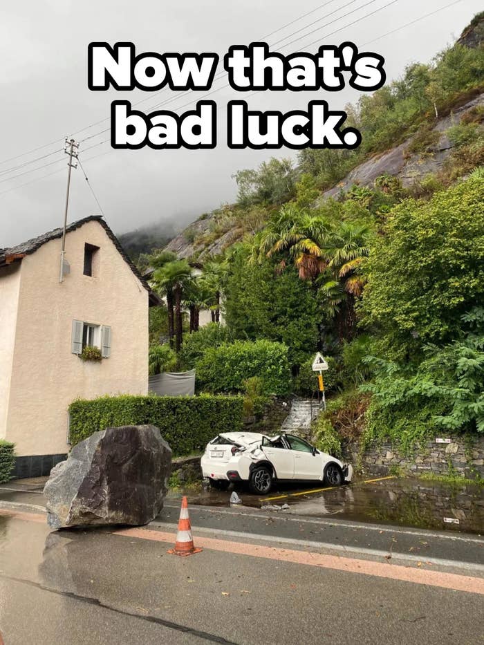 A large boulder has fallen onto a car parked beside a house on a street, causing significant damage to the car. No people are visible