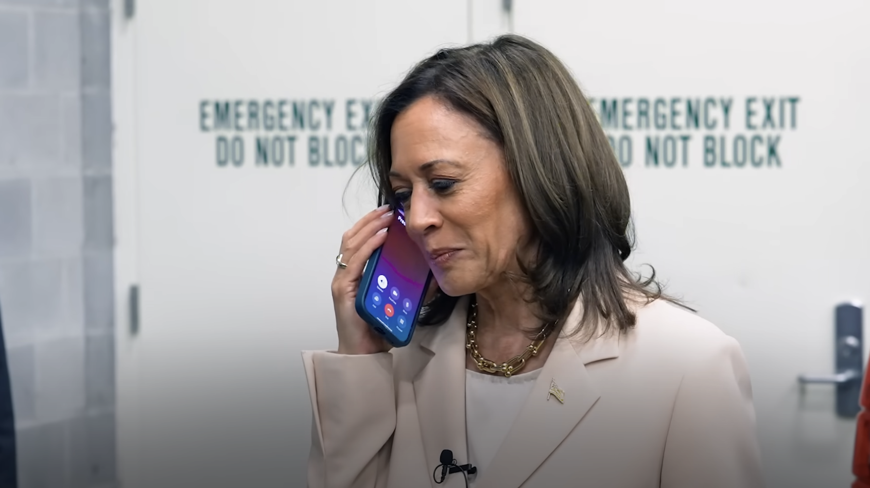 Kamala Harris smiling and holding a phone to her ear, standing in front of an emergency exit