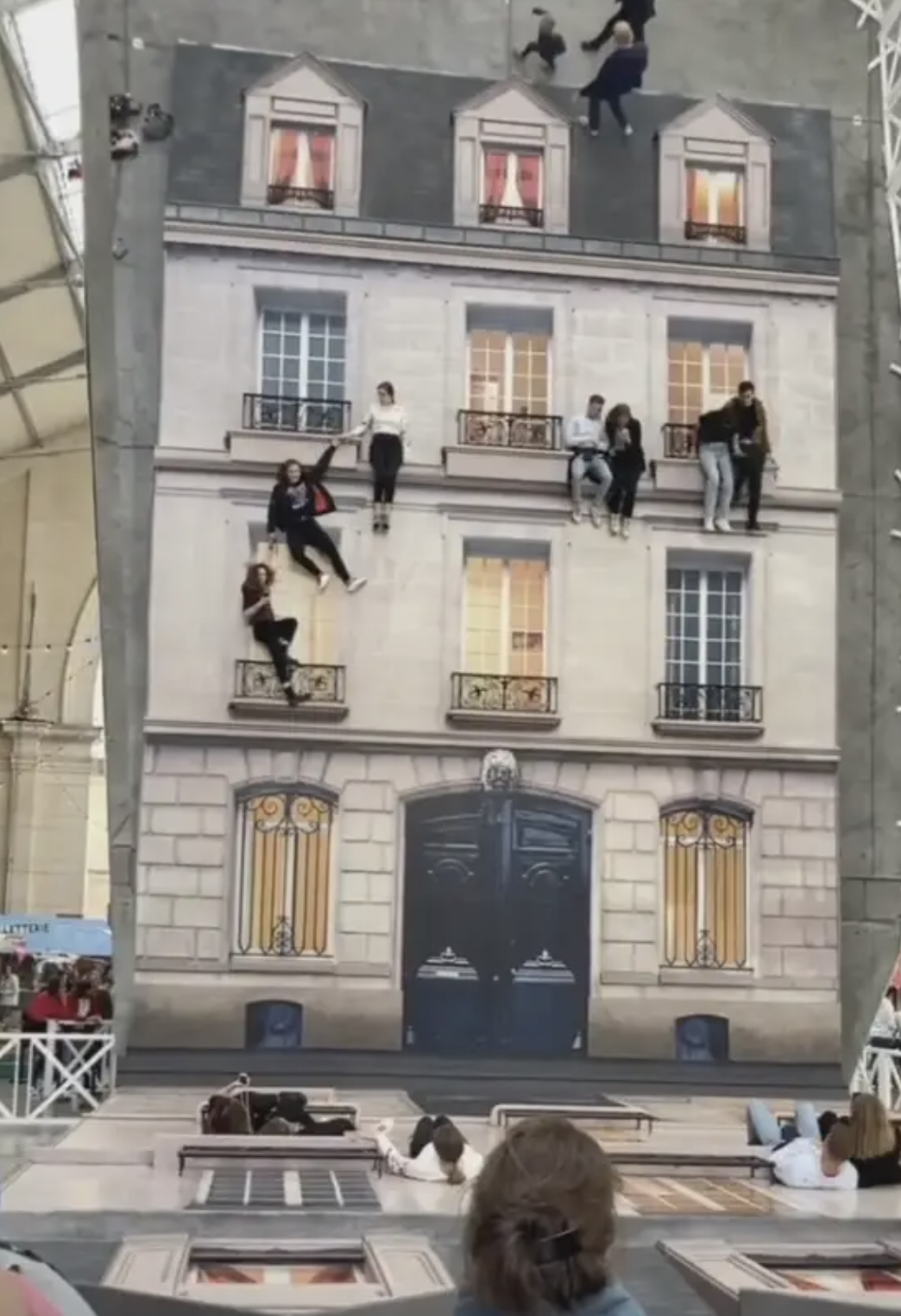 People appear to be climbing and hanging onto a large mural resembling the facade of a building, while others lay on the floor below observing