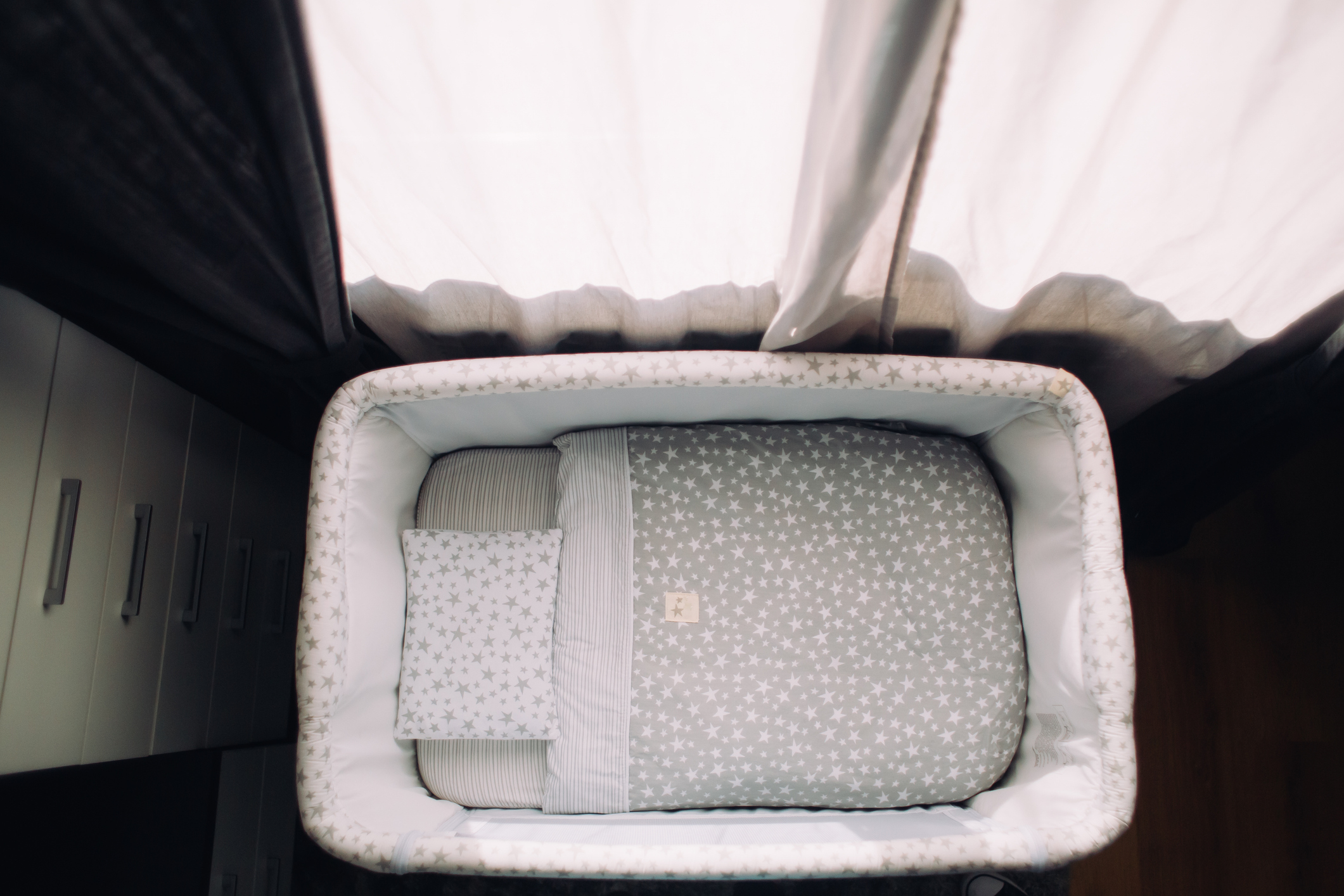 A cozy baby crib with a star-patterned mattress and pillow, placed under a window with sheer curtains, viewed from above. No persons in the image