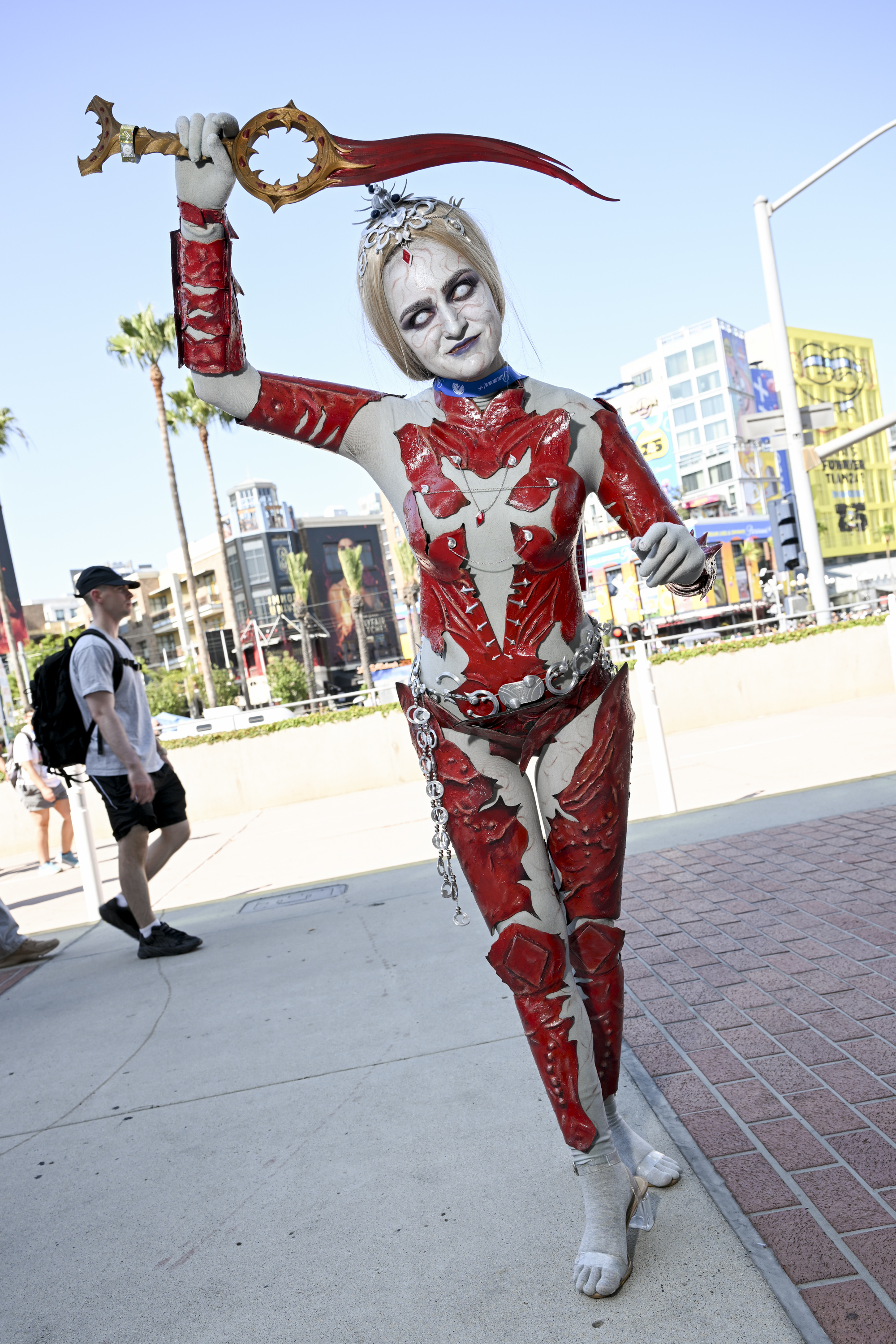 Person dressed as a character in red and white intricate costume, holding a saber aloft at an outdoor event. Other people are walking in the background