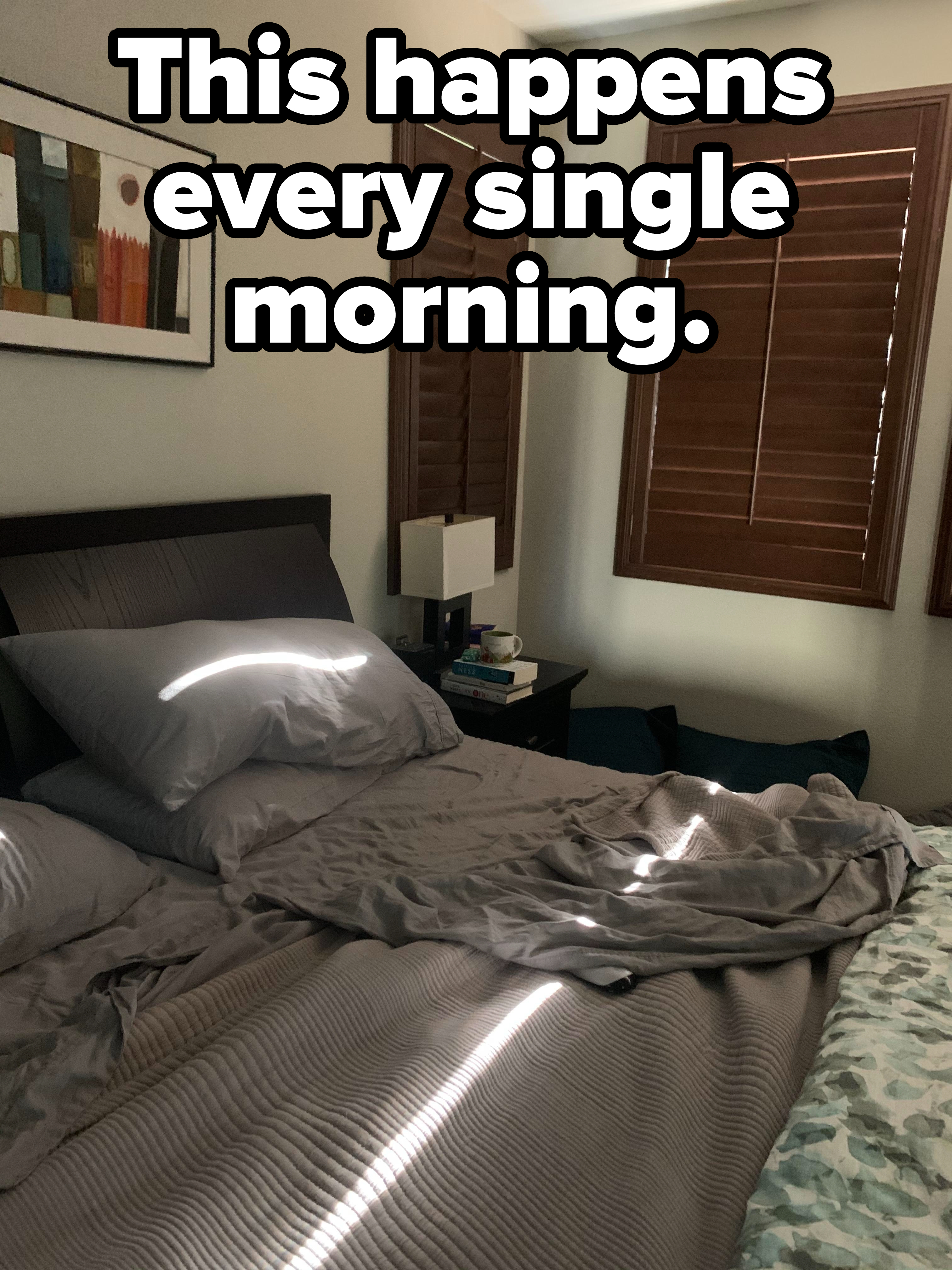A neatly made bed with gray bedding is bathed in sunlight streaming through closed wooden shutters. A nightstand beside the bed holds books and a lamp