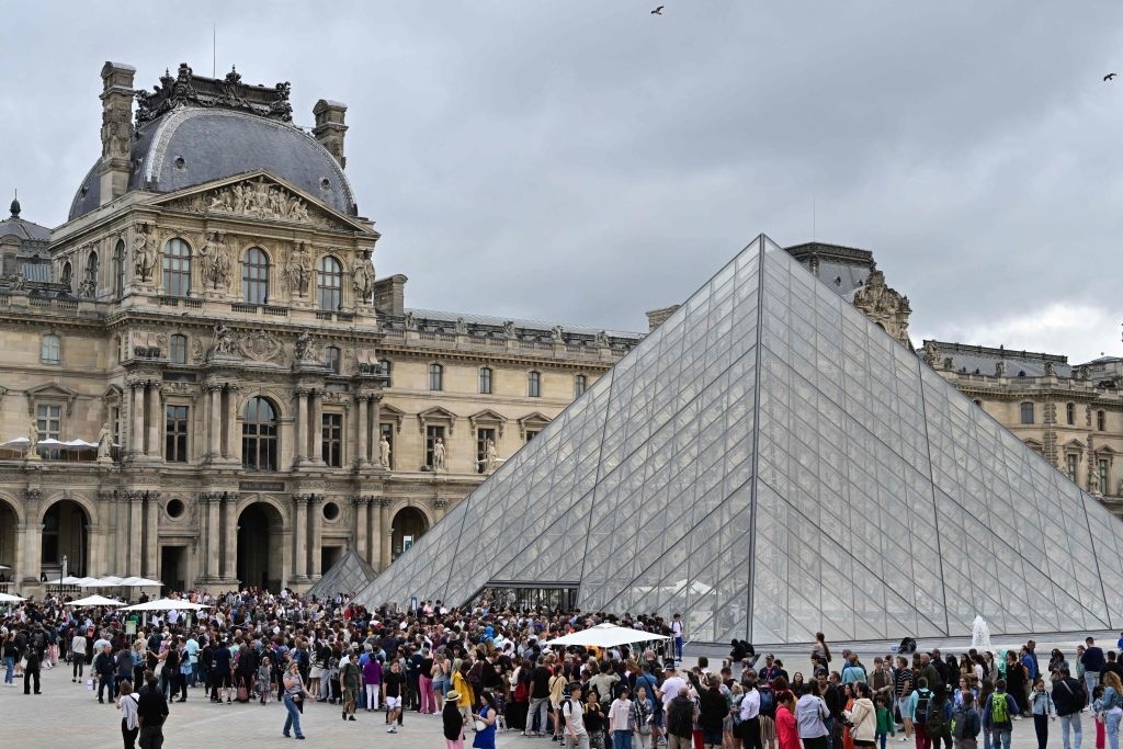 Crowds gather outside the glass pyramid entrance of the Louvre Museum in Paris, with the historic building in the background