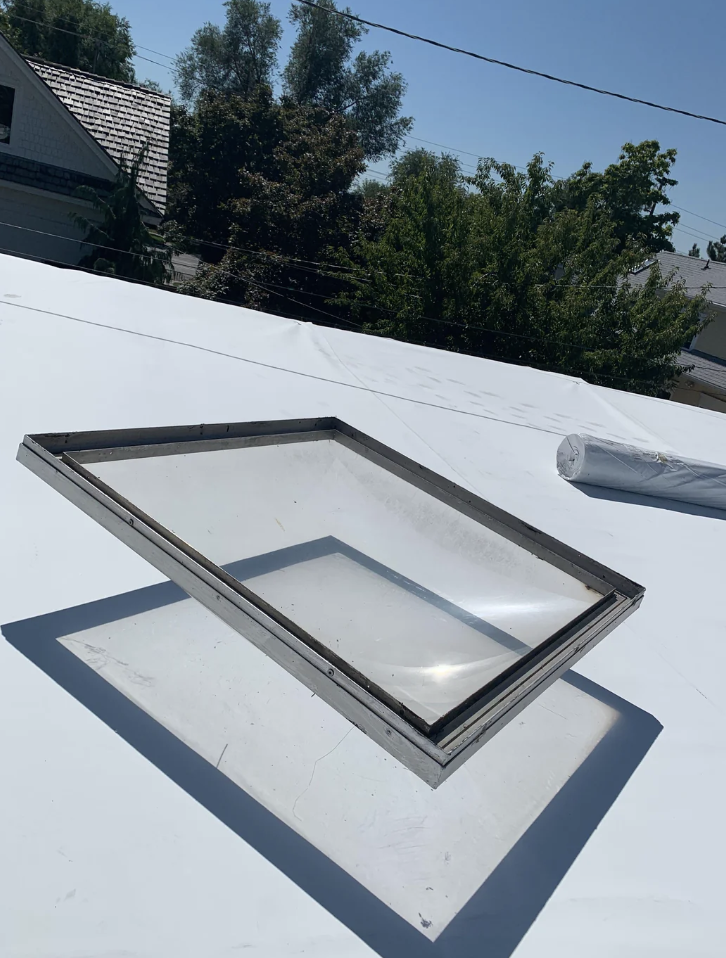 A rectangular skylight is laid flat on a white rooftop with some trees and a house in the background