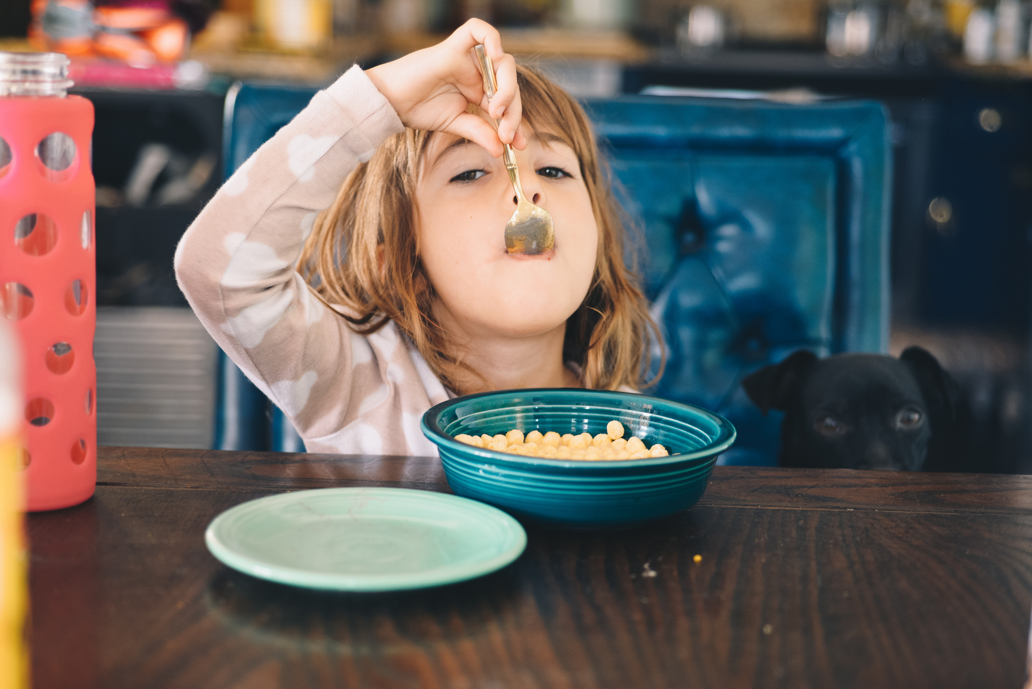 A child eats cereal from a blue bowl while a black dog peeks from behind the table