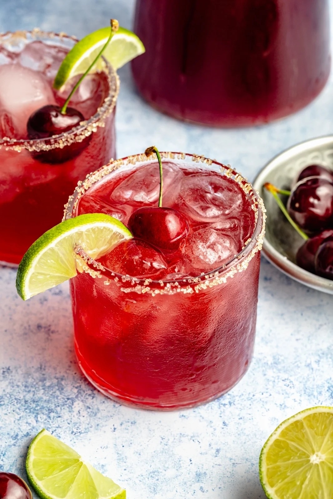 Two glasses of a red cocktail with ice, garnished with lime slices and cherries, on a blue surface with additional lime and cherries nearby