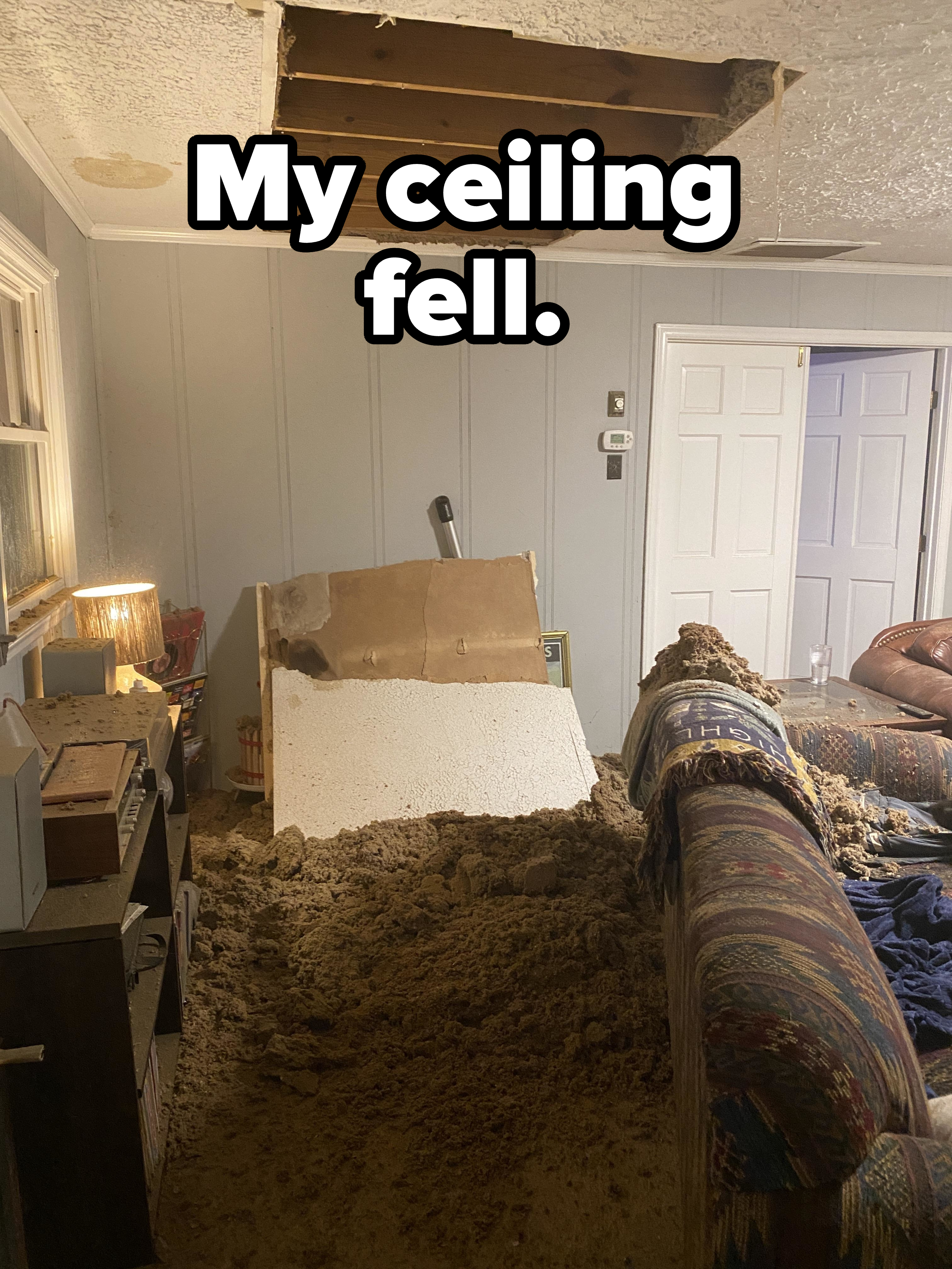 Living room with a large hole in the ceiling, drywall and insulation collapsed onto a fallen couch. The rest of the room appears cluttered but undamaged