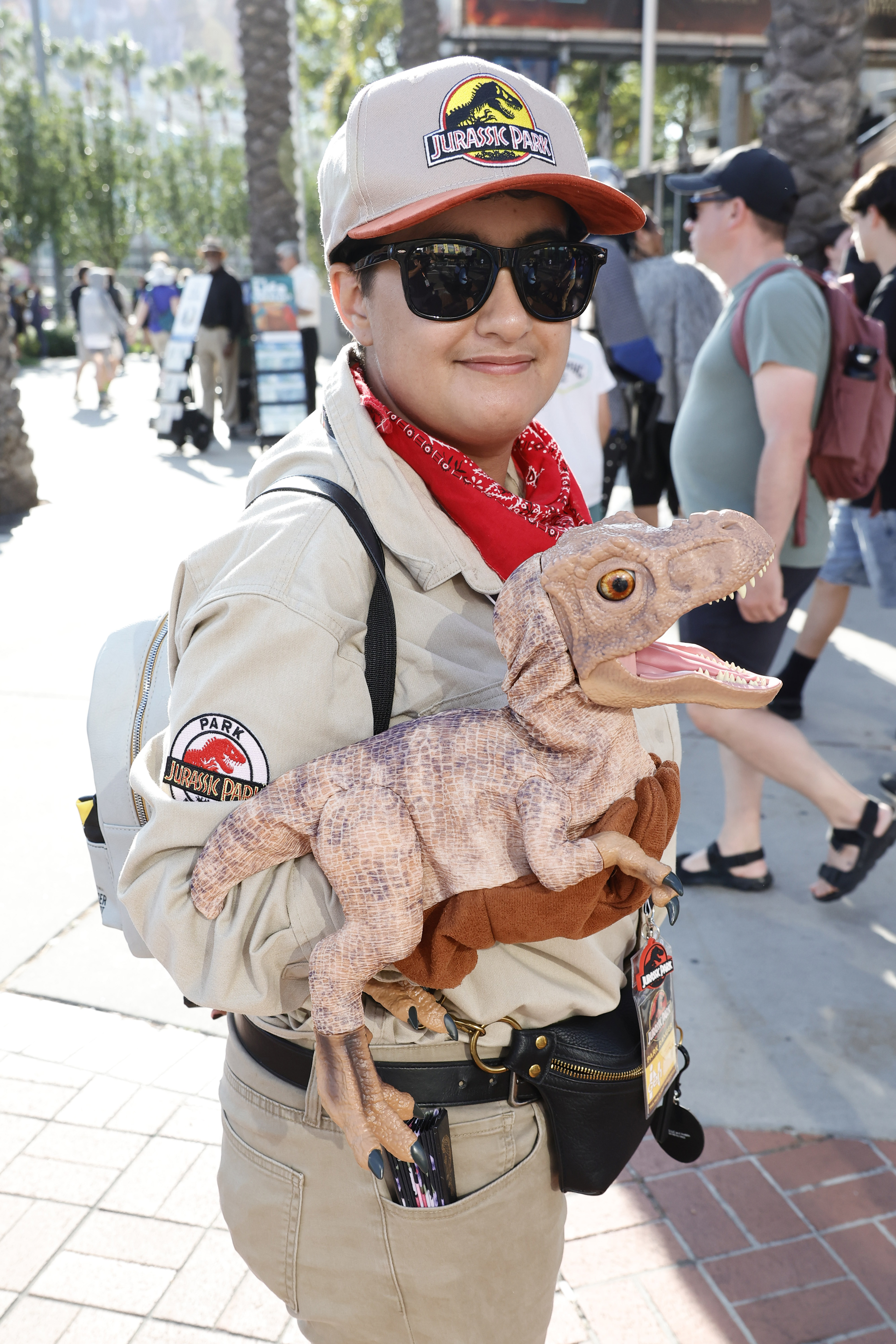 Person holding a dinosaur toy, dressed in a Jurassic Park-themed outfit with sunglasses, tan hat, and red bandana, standing outdoors among a crowd
