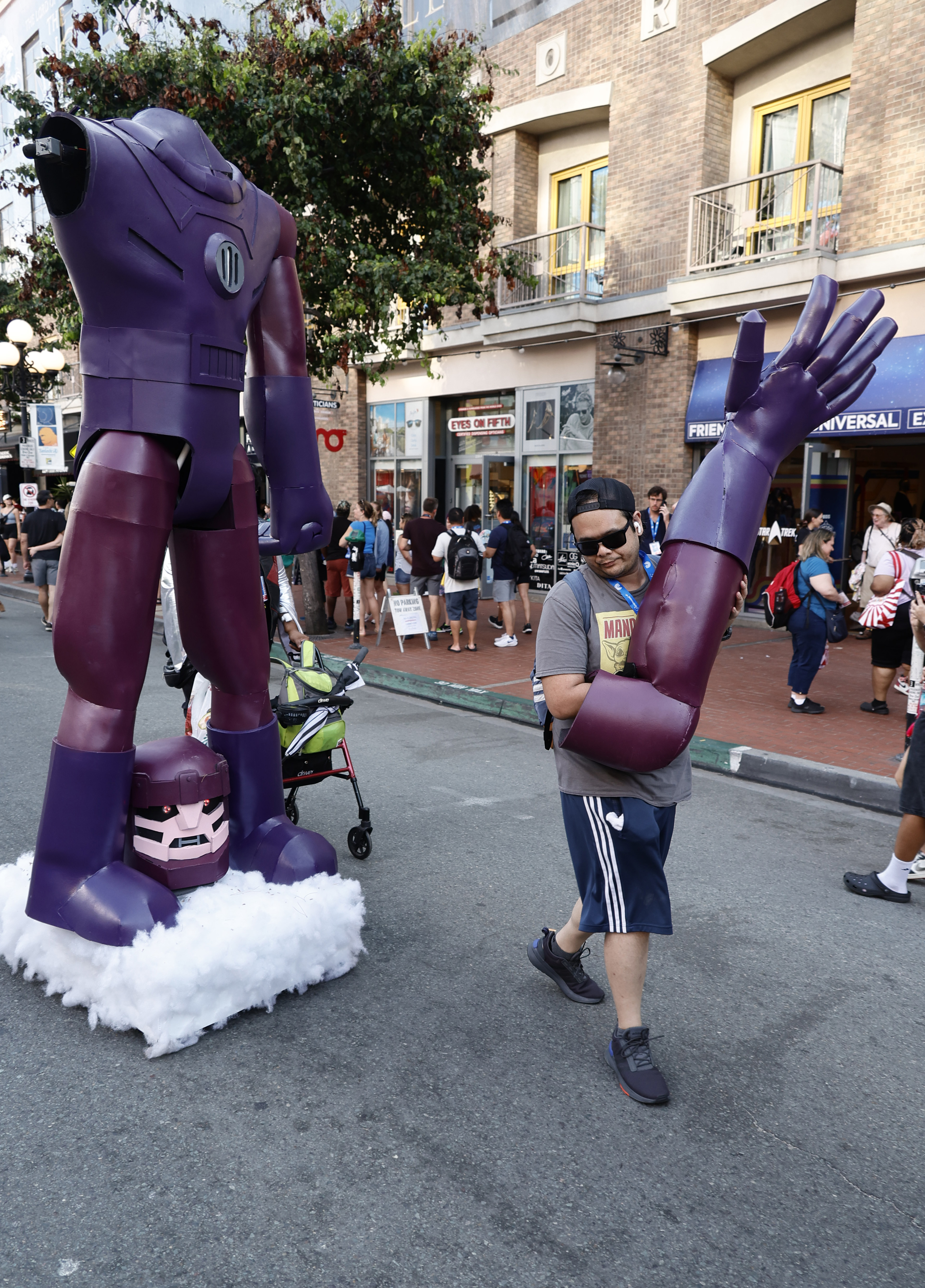 A person in a t-shirt and shorts carrying a giant purple robot arm from a large robot costume in a busy street with passersby
