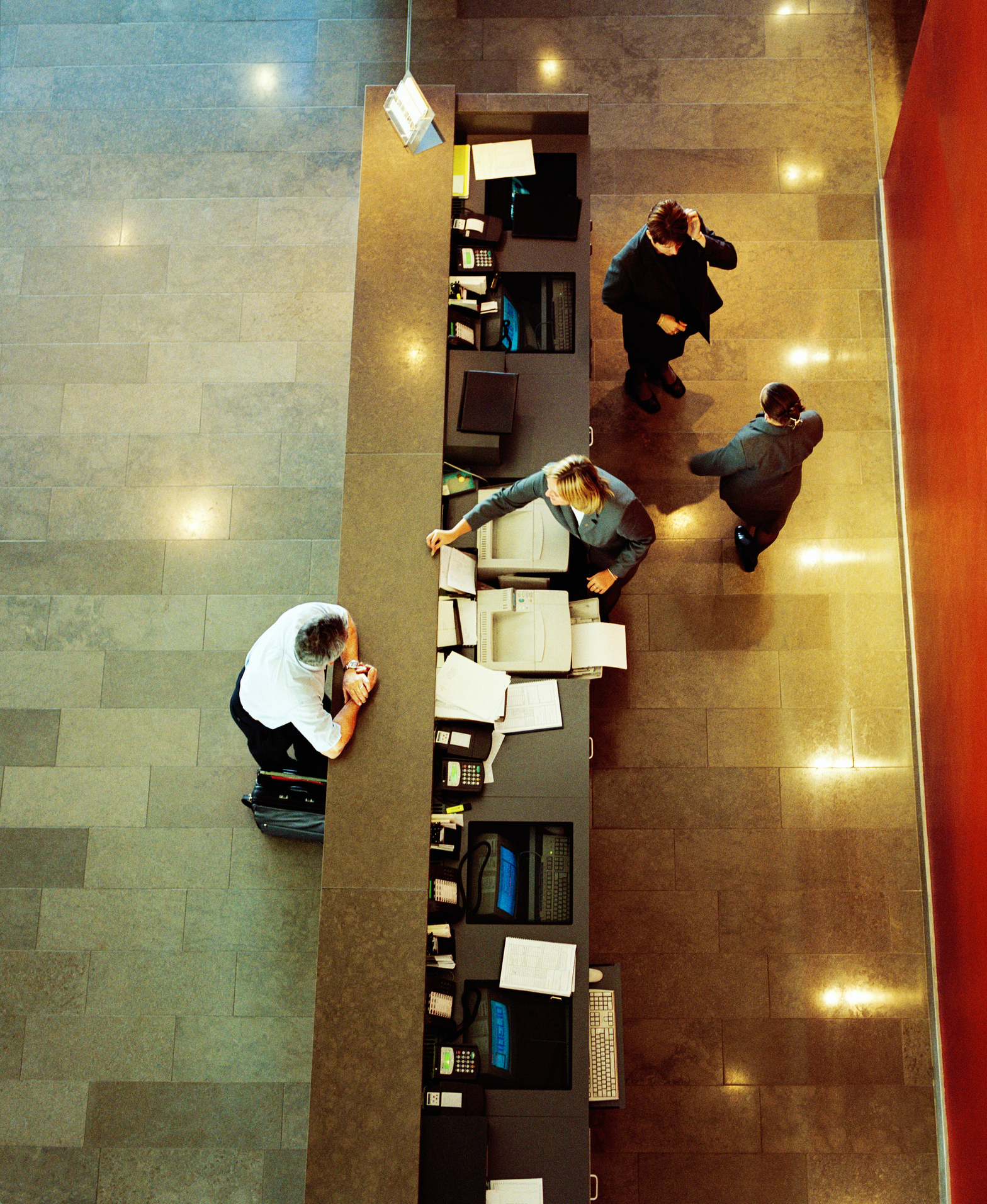 Aerial view of four people at a reception desk, including a receptionist attending to a visitor and two other individuals standing and interacting nearby
