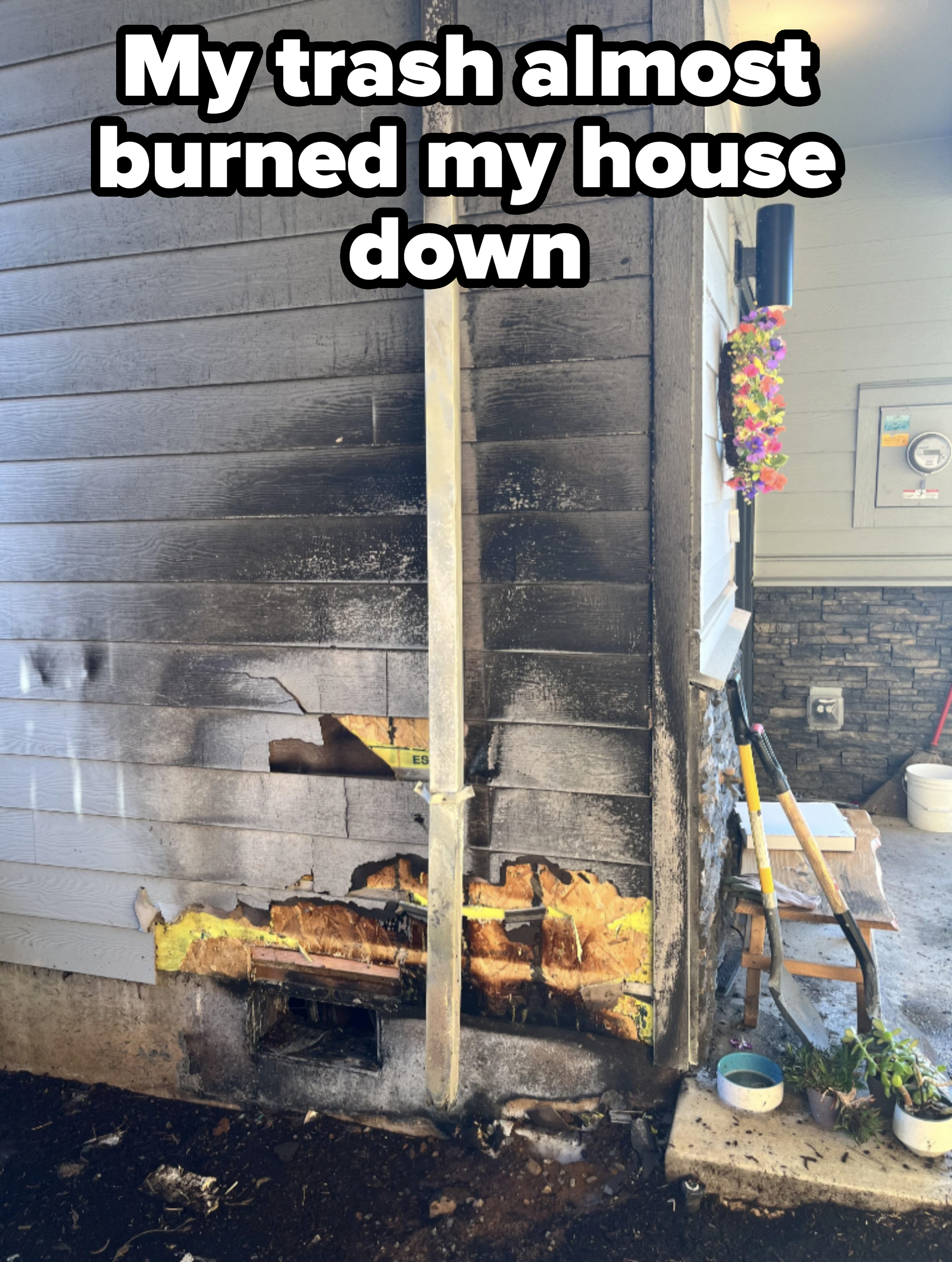 A damaged exterior house corner with significant burn marks, charred siding, and burnt insulation. A tall decorative flower garland is visible on the wall nearby