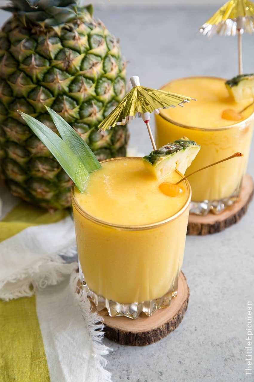 Two pineapple cocktails with garnish on wooden coasters next to a whole pineapple and a cloth napkin