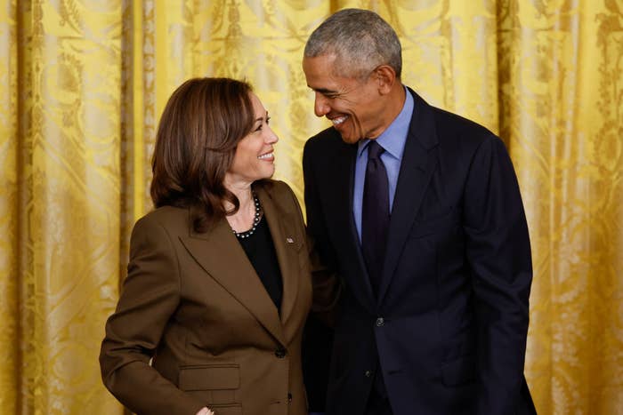 Kamala Harris and Barack Obama smiling at each other, standing in front of a decorative curtain. Harris is in a suit, and Obama is in a suit with a tie