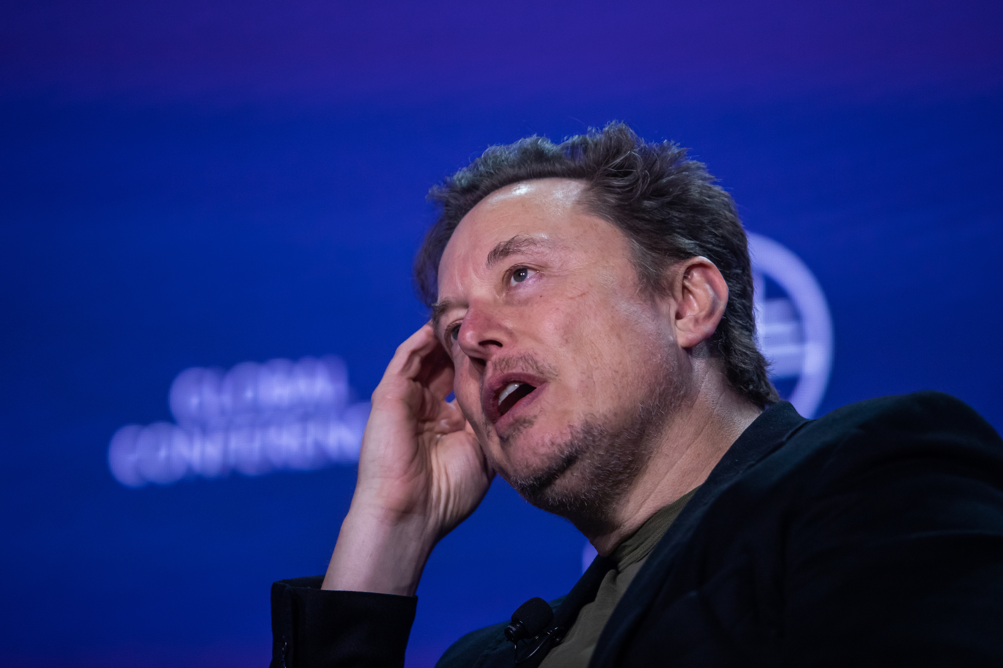 Elon Musk speaks at the Global Conference, holding his hand to his head while wearing a dark jacket