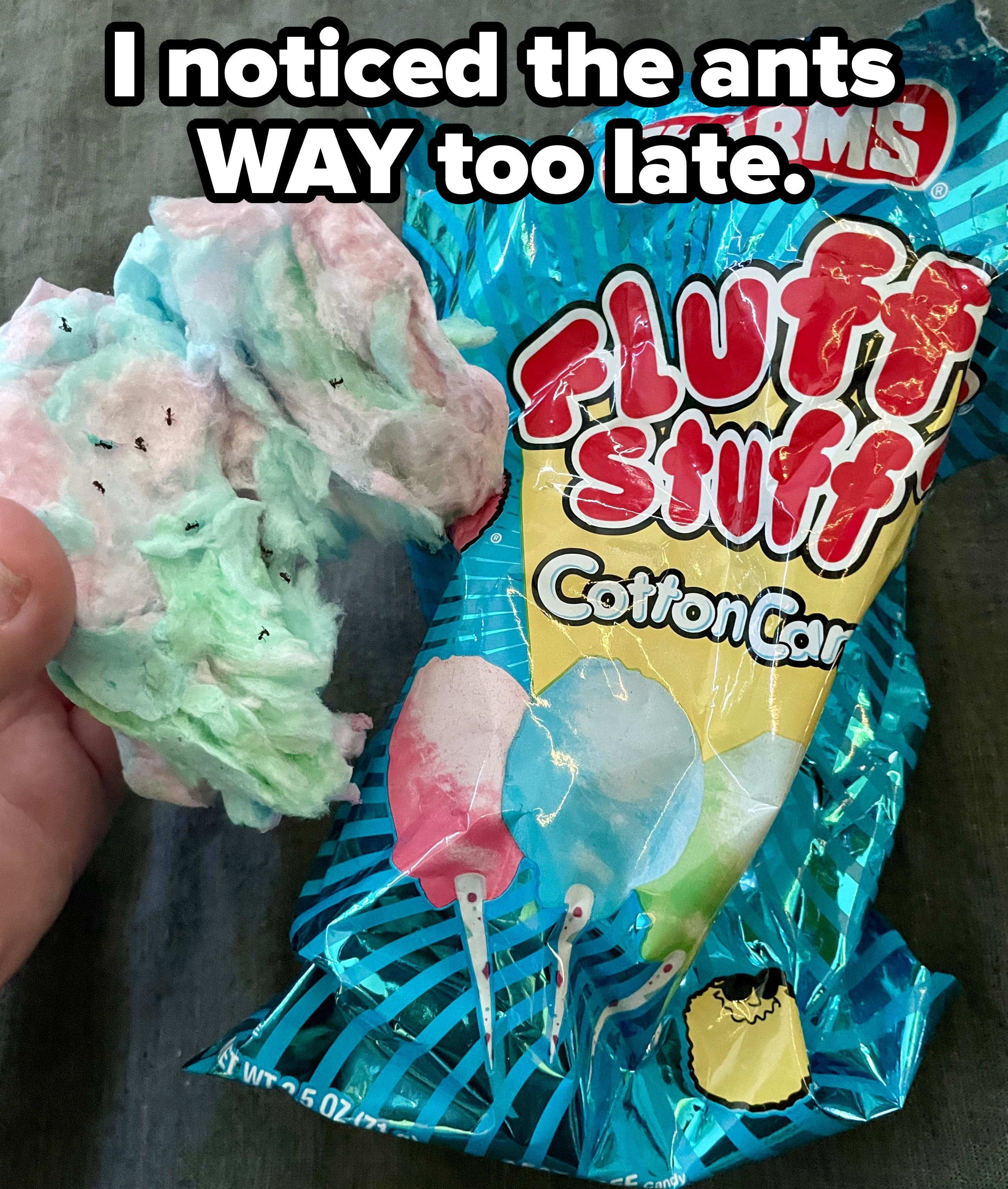 A hand holds a piece of pink and green cotton candy next to an opened Charms Fluffy Stuff Cotton Candy bag