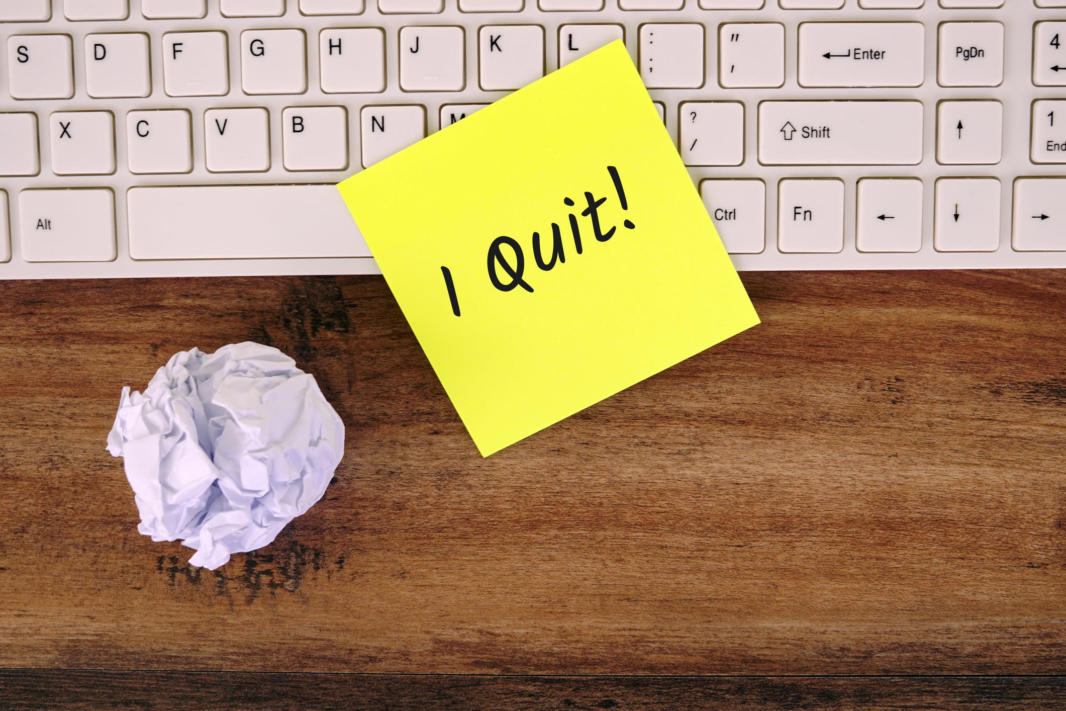 A yellow sticky note with &quot;I Quit!&quot; written on it is placed on a wooden desk next to a crumpled piece of paper and a keyboard