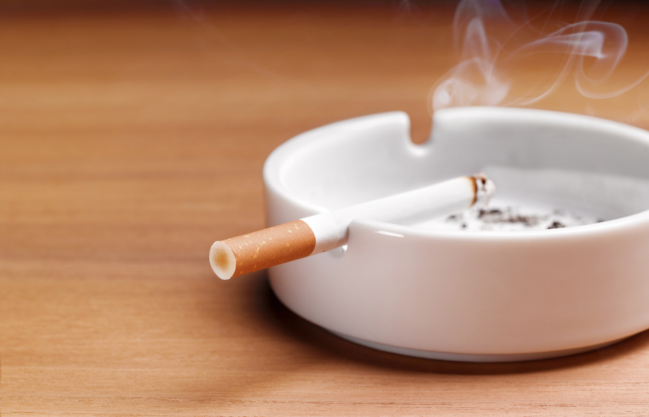 A lit cigarette is resting on the edge of a white ashtray, with smoke rising from its tip