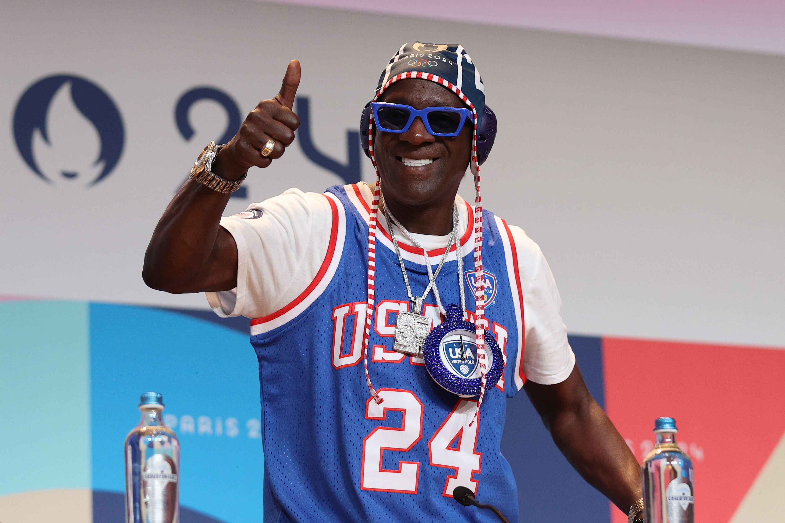 Flavor Flav wears a blue USA basketball jersey and oversized clock necklace, giving a thumbs up at a press event