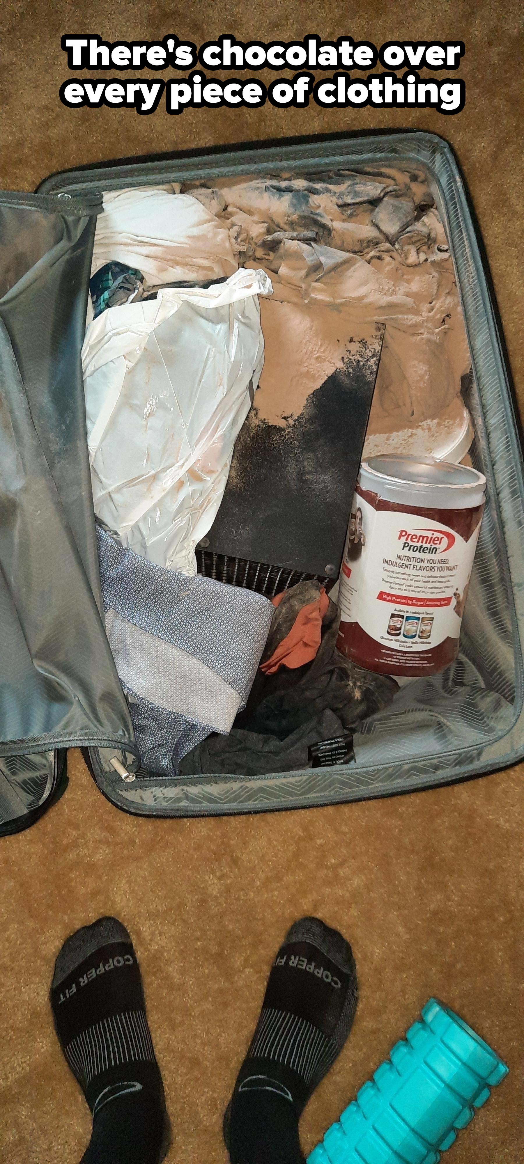 Open suitcase on a carpet; contents include spilled protein powder, clothing items, a small canister, and other miscellaneous items. Visible feet in socks at the bottom