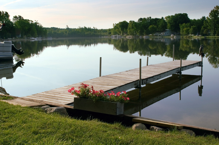 A peaceful lakeside scene with a dock jutting into calm water. A pot of flowers is placed near the dock. Trees and houses are visible in the background