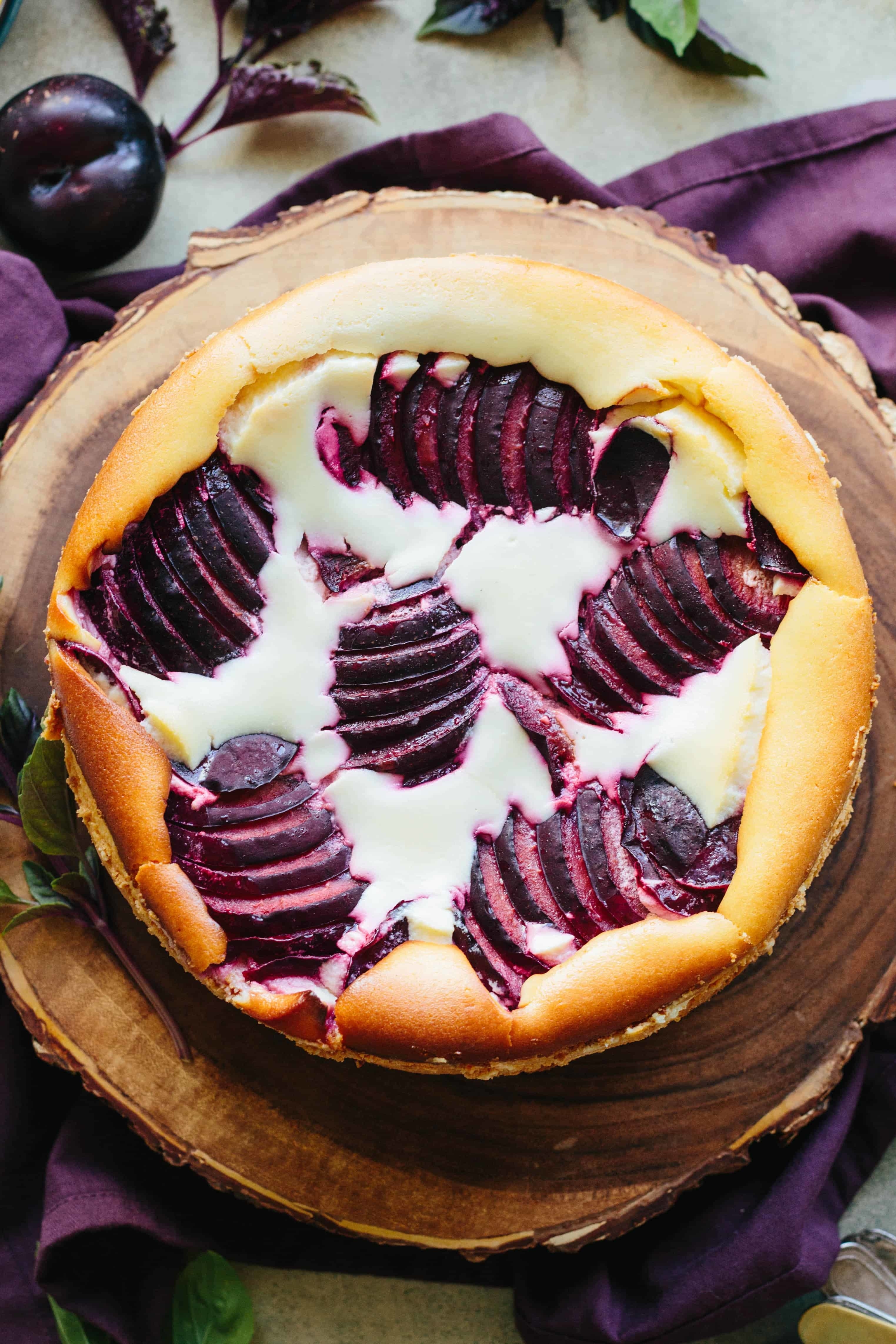 Cheesecake with sliced plums on top, placed on a round wooden board. Purple cloth and plums are in the background