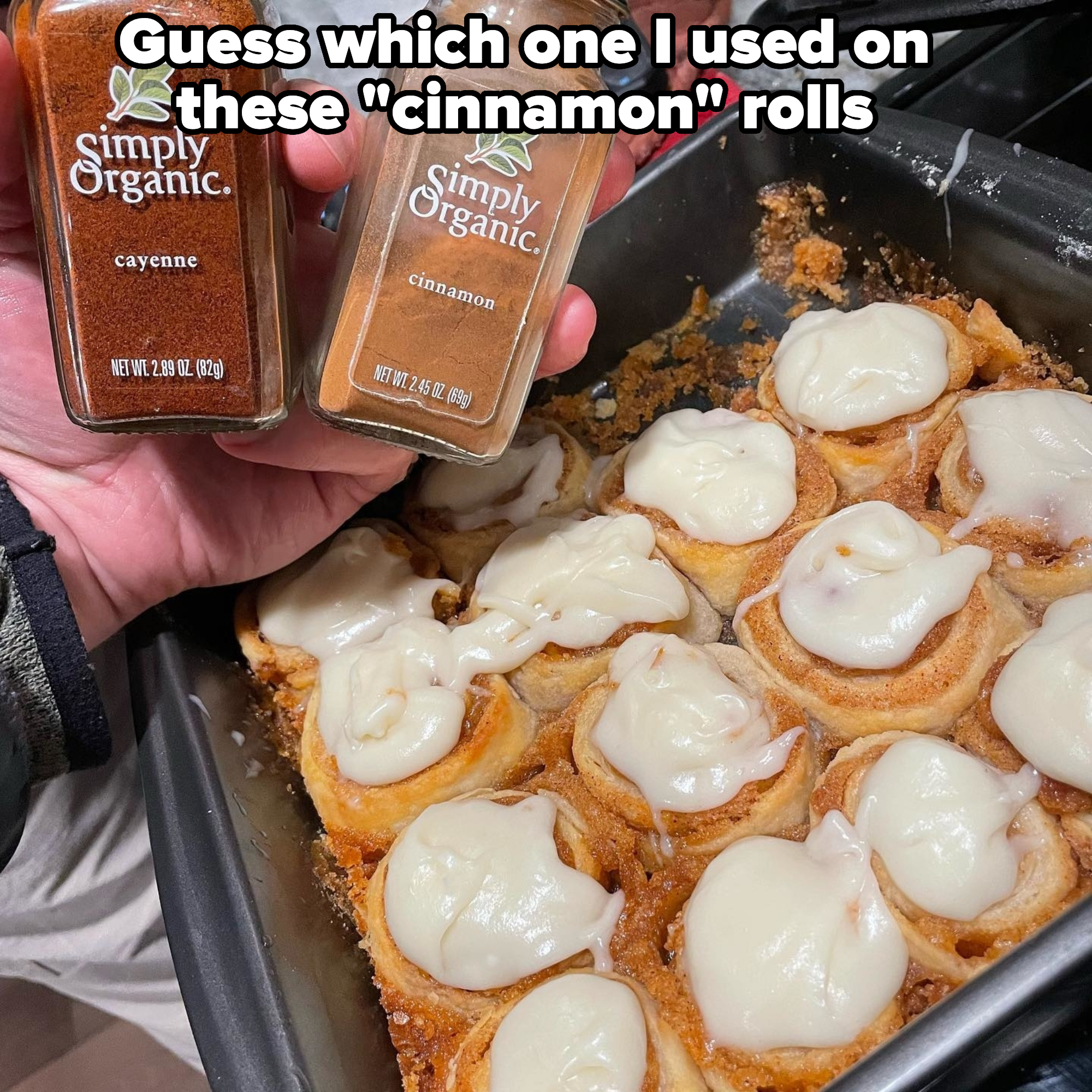 A close-up of a tray of cinnamon rolls with cream cheese frosting. A hand holds jars of Simply Organic cayenne and cinnamon spices above the tray