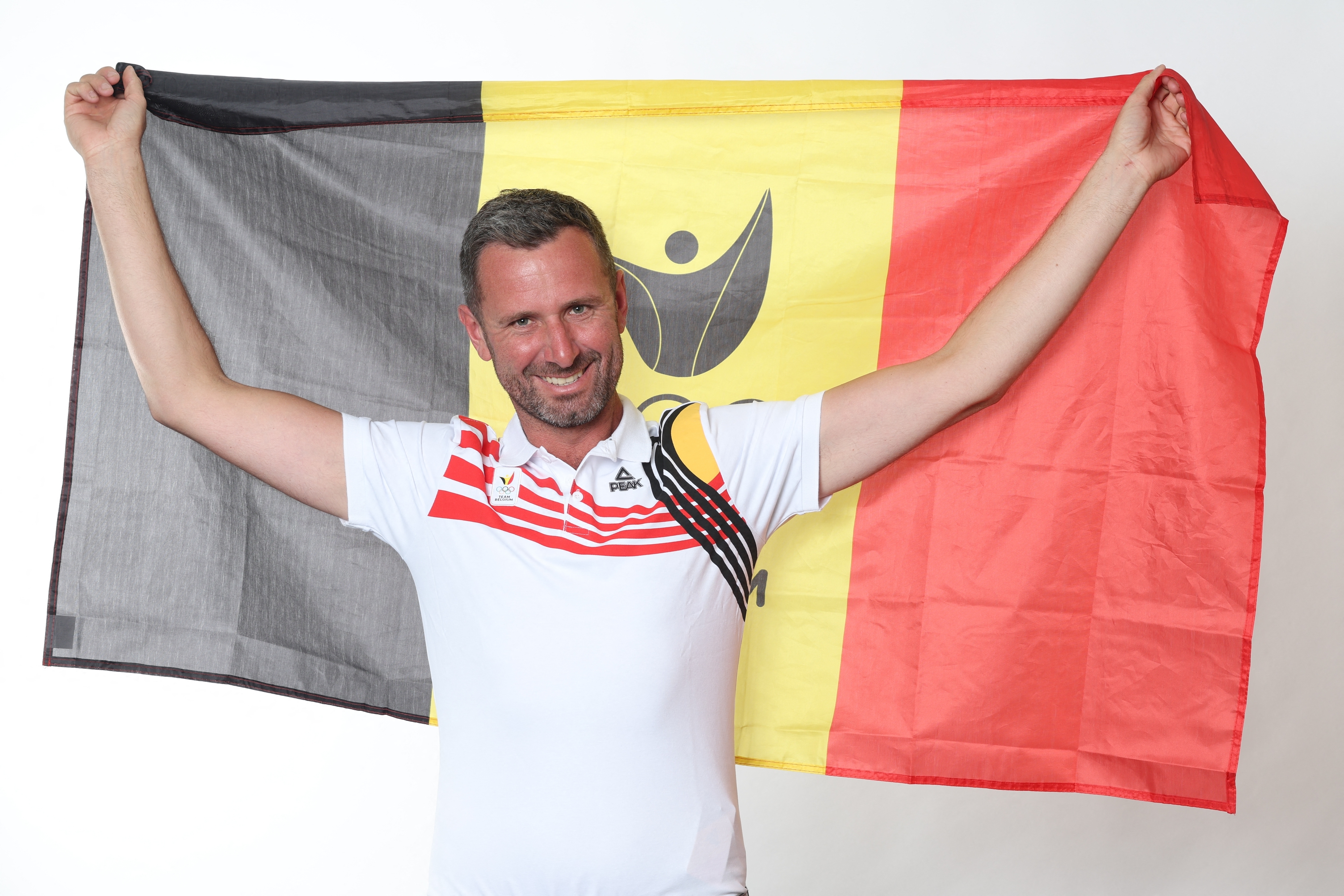 A man smiles and holds a Belgium flag behind him, dressed in a sports-themed white shirt with red and black accents