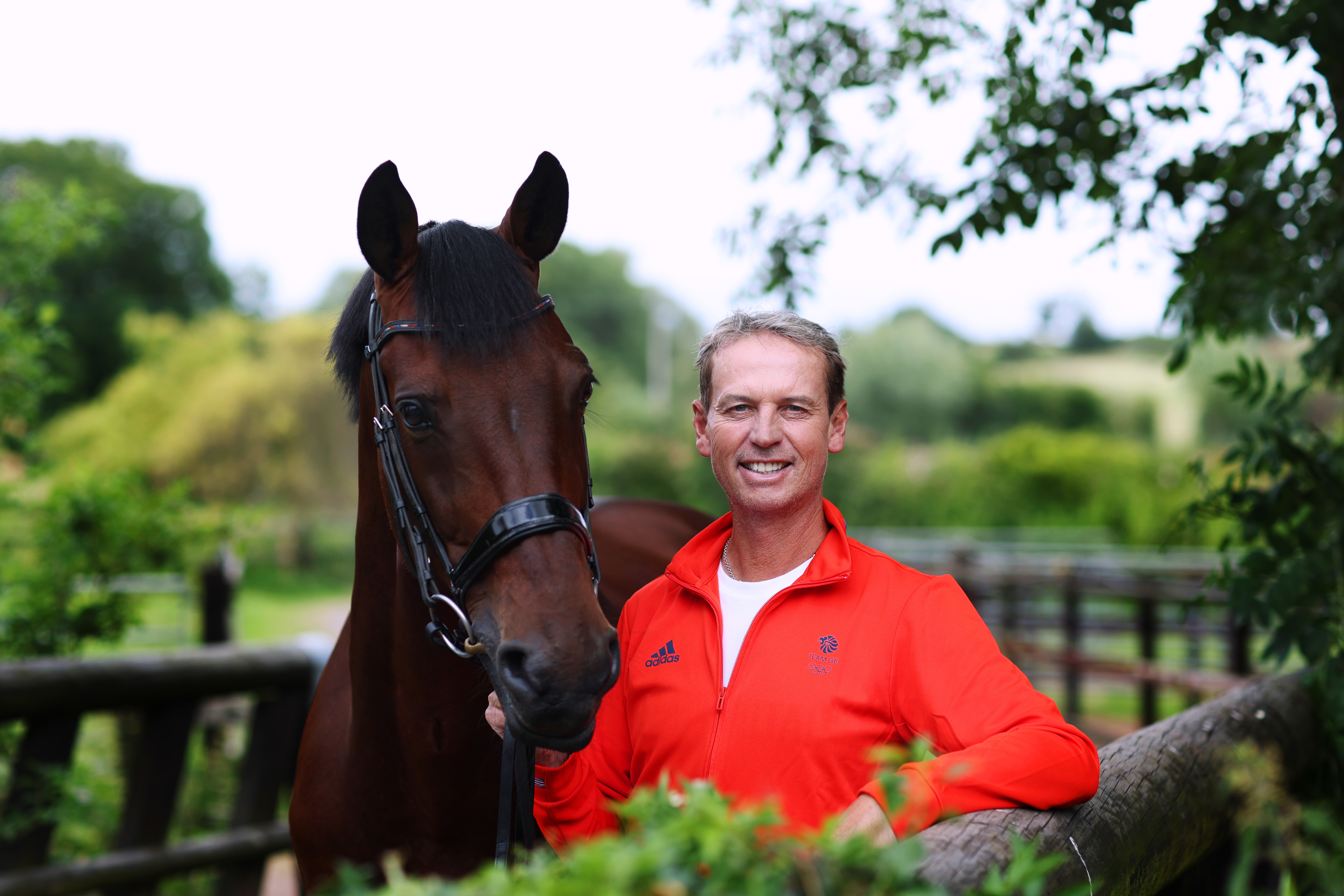 A man in a red Adidas zip-up jacket stands next to a horse in a lush, green outdoor setting, both looking at the camera