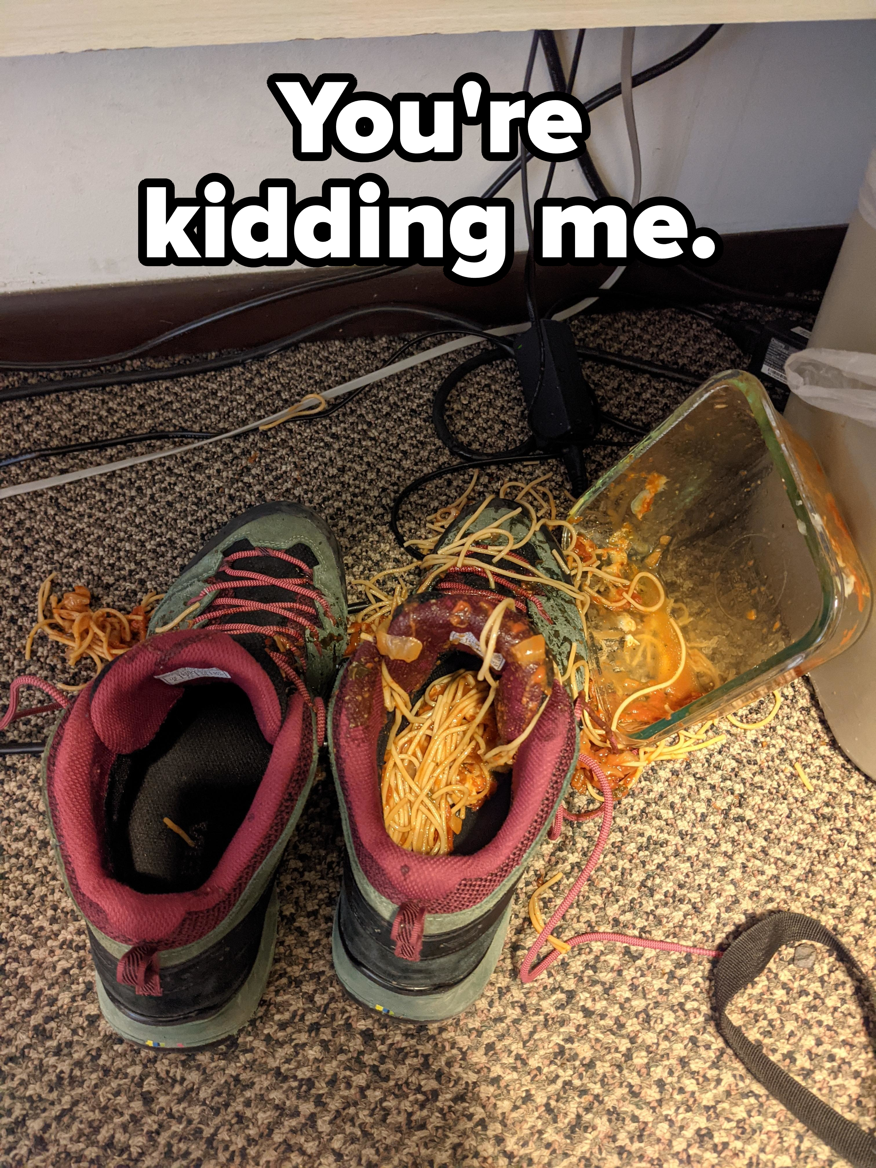 Two hiking boots filled with spilled spaghetti and sauce under a desk with tangled wires
