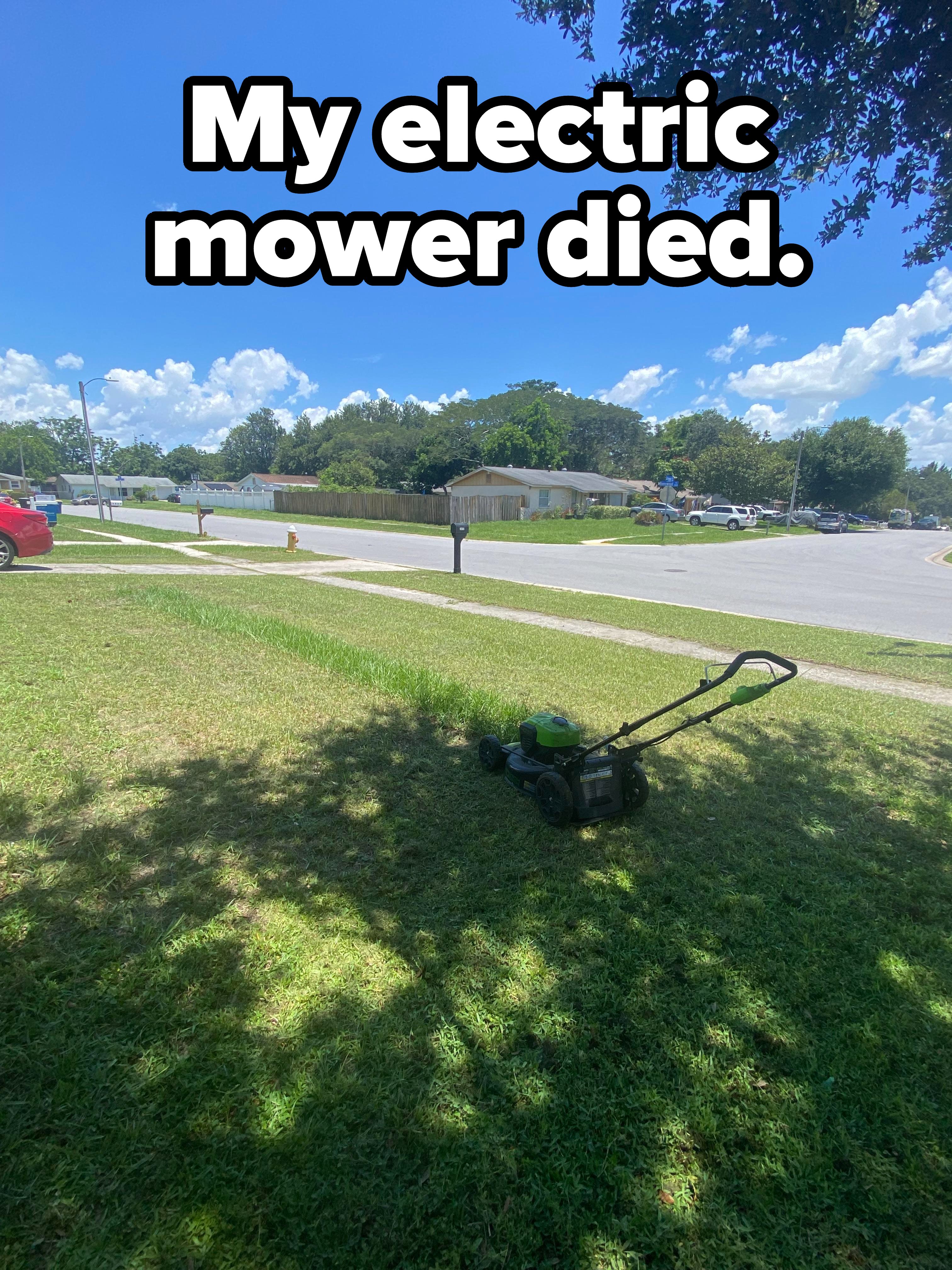 A lawnmower sits in the middle of a grassy lawn, with a residential street and houses visible in the background under a clear sky. No people are present
