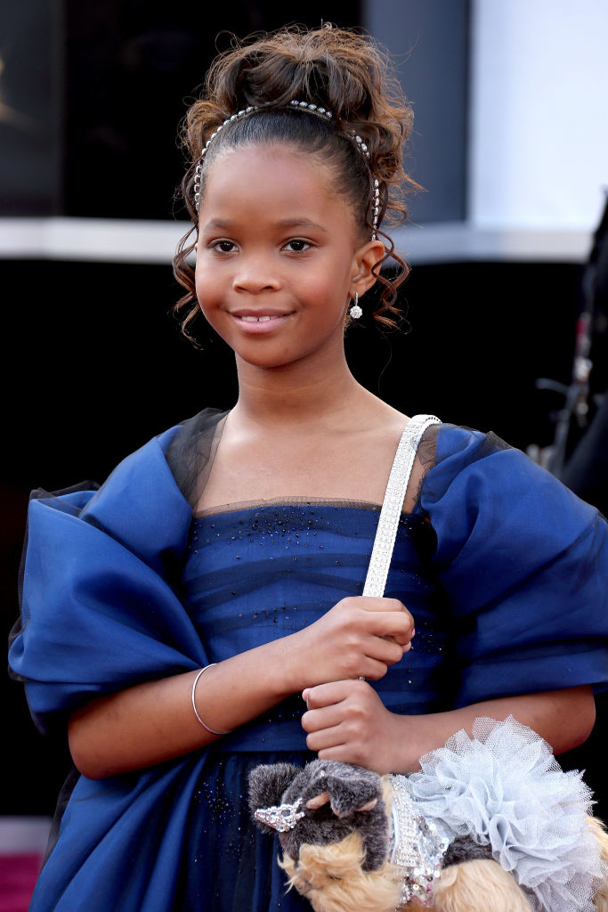 Quvenzhané Wallis on the red carpet wearing an elegant dress with a sparkly headband and holding a plush puppy handbag