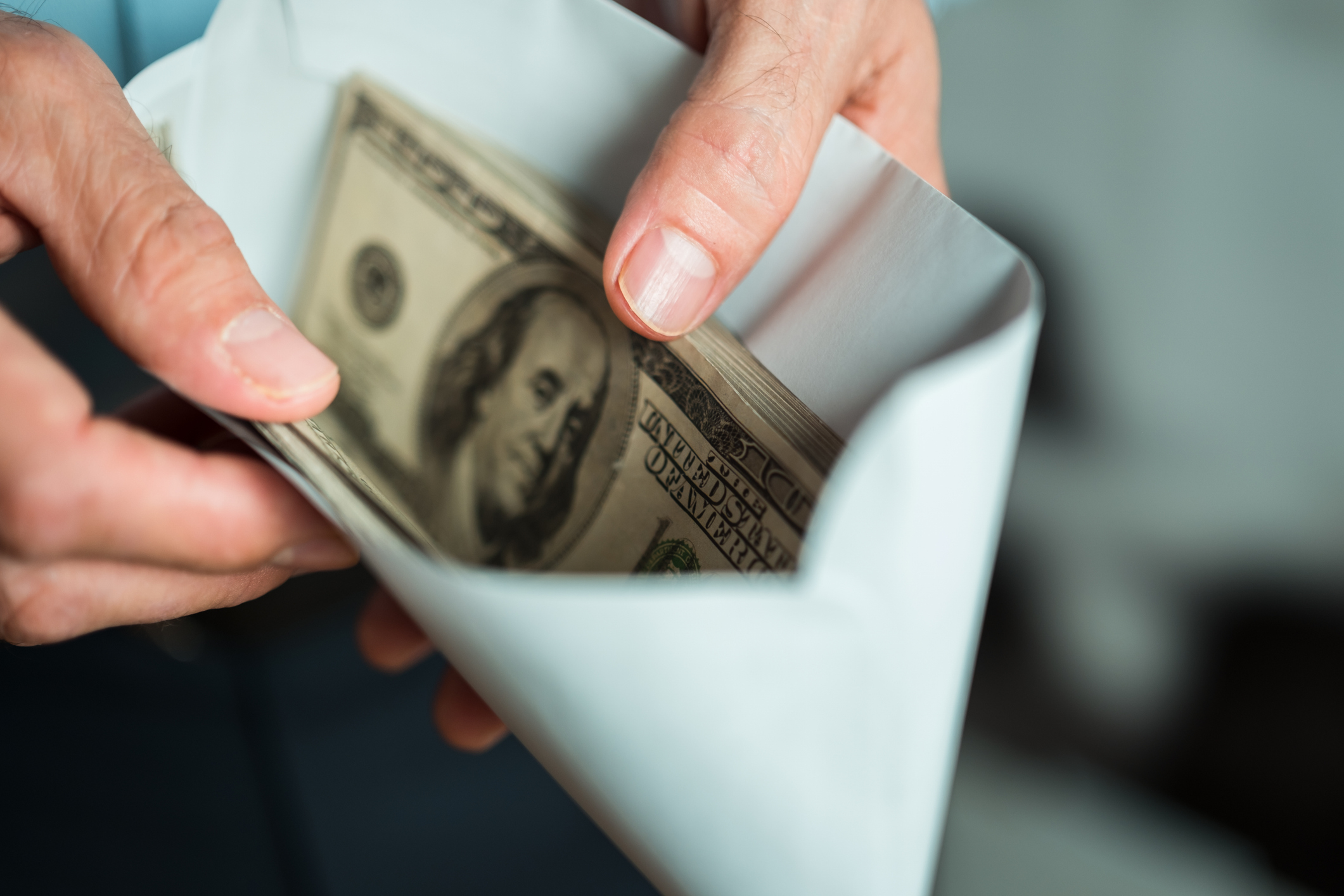 Hands holding a white envelope with a stack of hundred-dollar bills partially visible inside