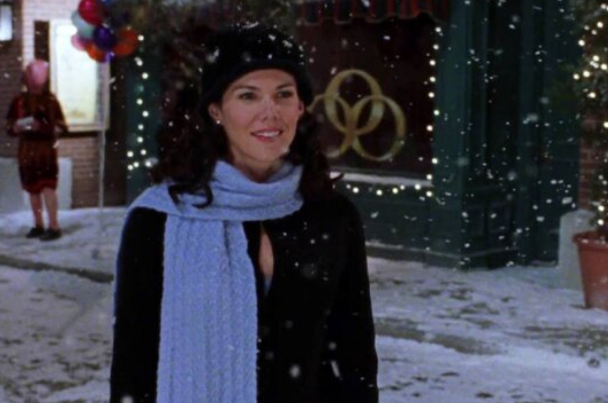Lauren Graham stands in the snow wearing a winter hat, scarf, and coat, with festive lights and balloons in the background