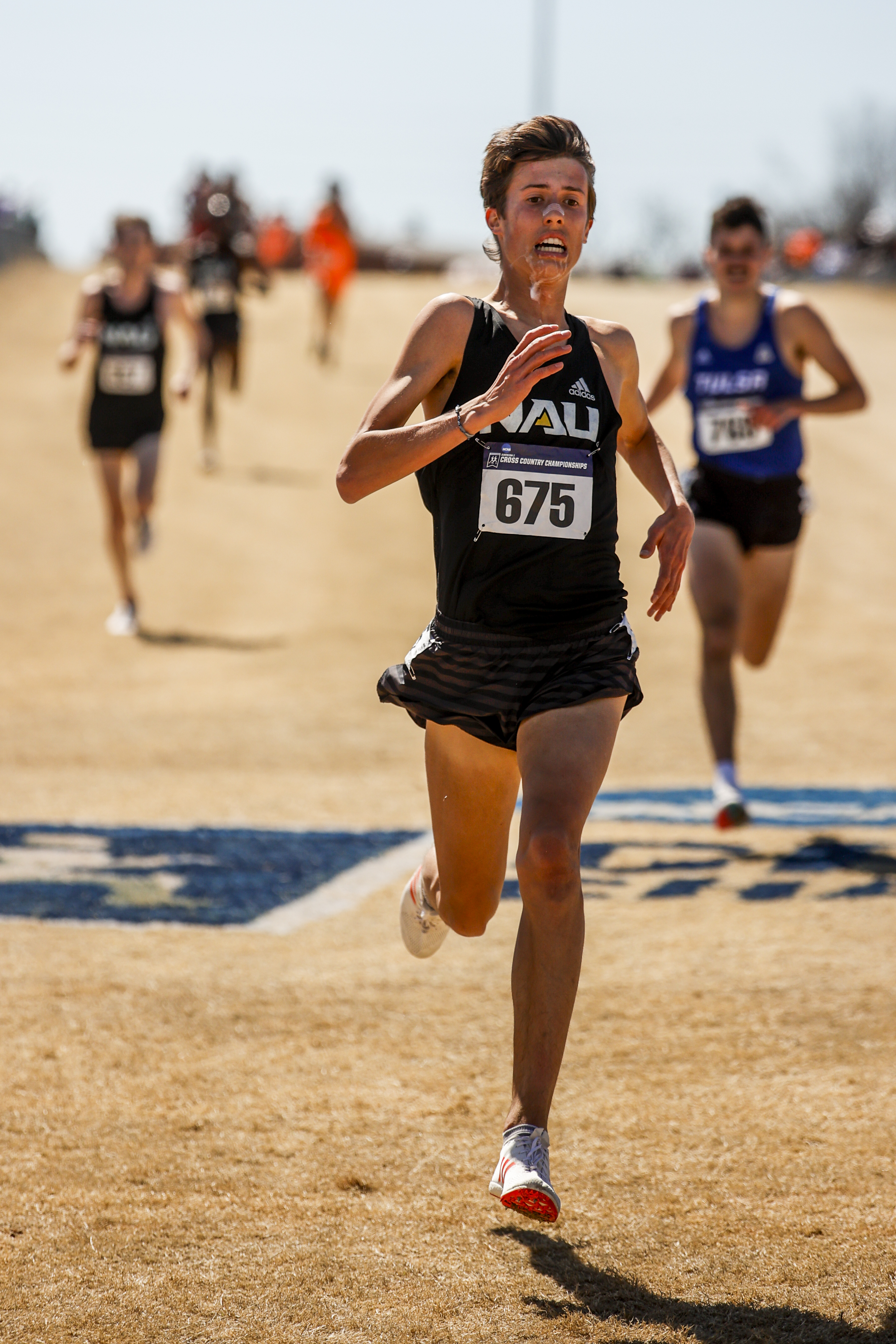 Runners are competing in a race, with a focus on one runner in a black uniform marked "NAU" and wearing bib number 675 in the foreground