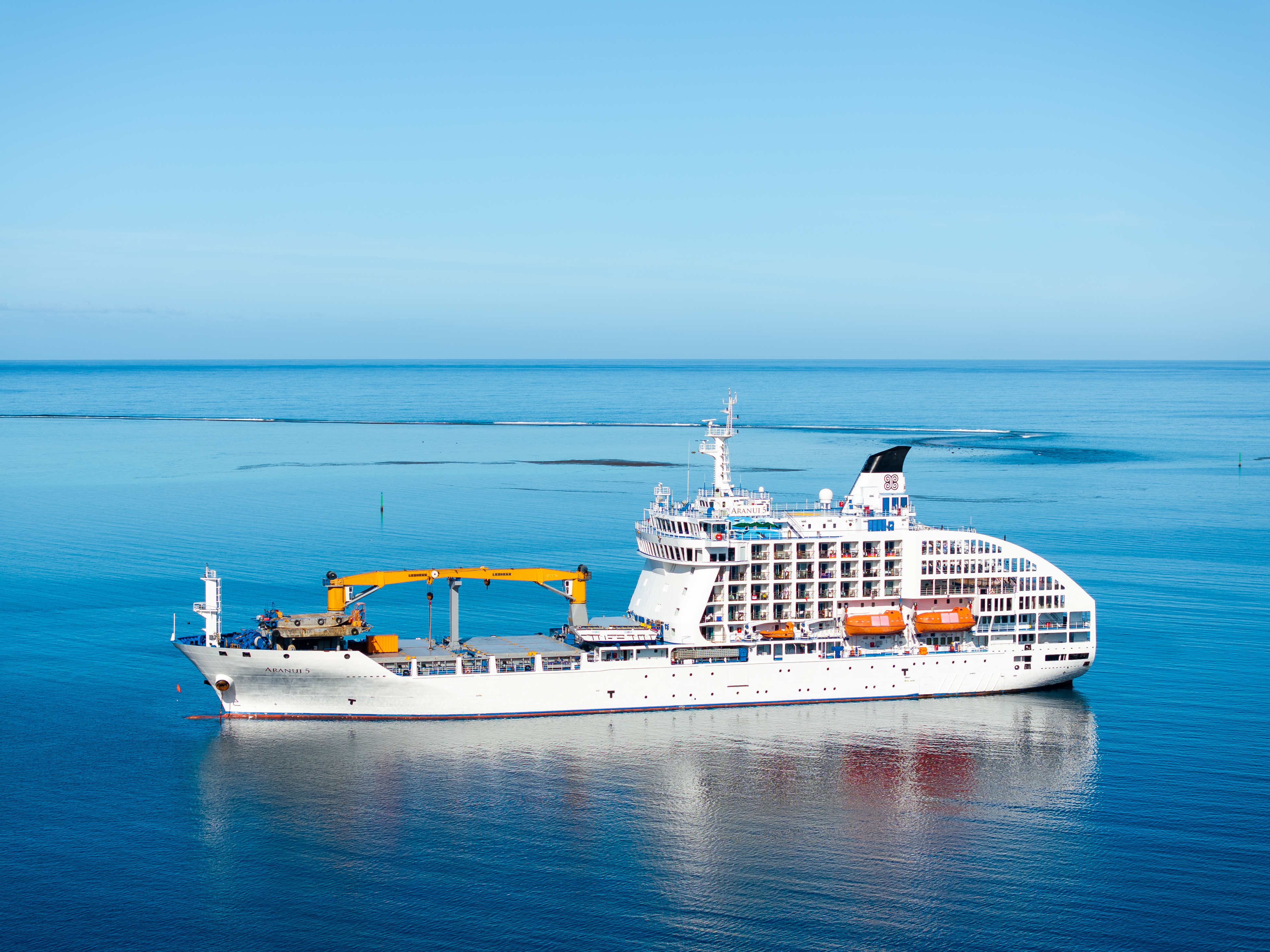 A large cruise ship sails on calm ocean waters under a clear sky. The ship's deck features yellow cranes and lifeboats