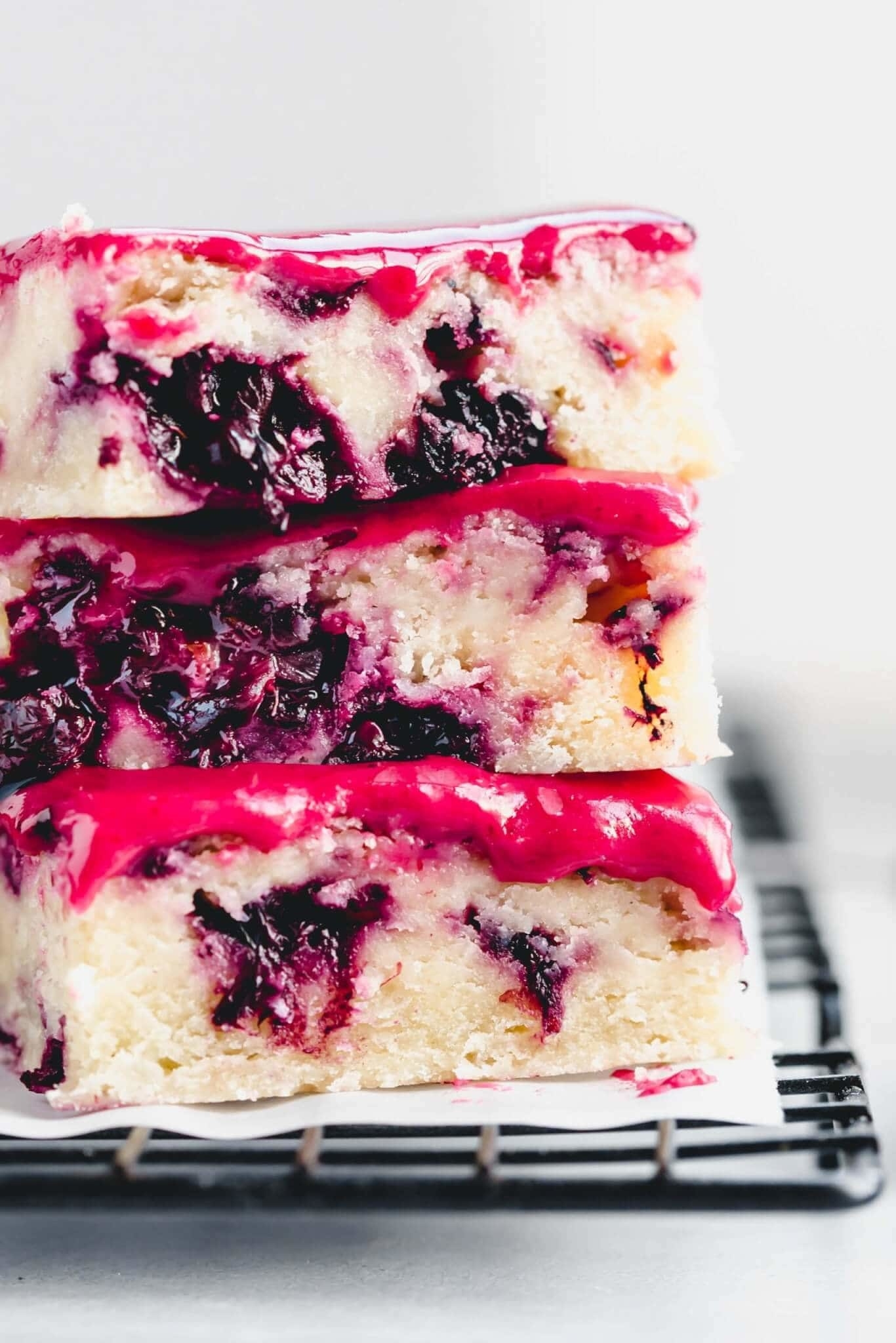 Three pieces of blackberry and lemon dessert bars stacked on a cooling rack. The bars feature a cream-colored base with purple-black fruit swirls and pink icing on top