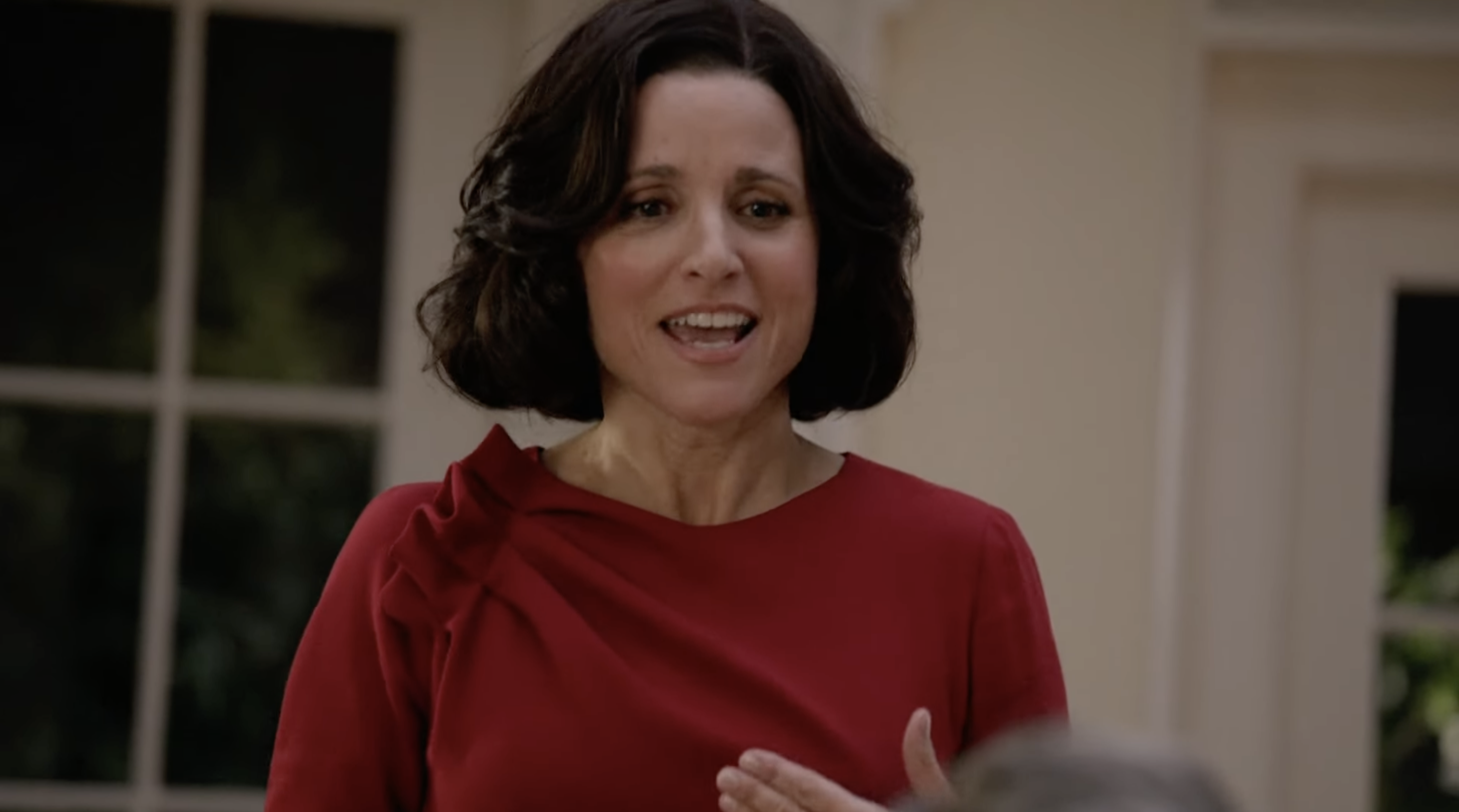 Julia Louis-Dreyfus, in a red dress, is speaking. She appears to be indoors