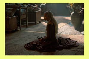 Rhaena from House of the Dragon with long white braids sits on the ground in a dimly lit room, holding an object. They wear a flowing, dark gown
