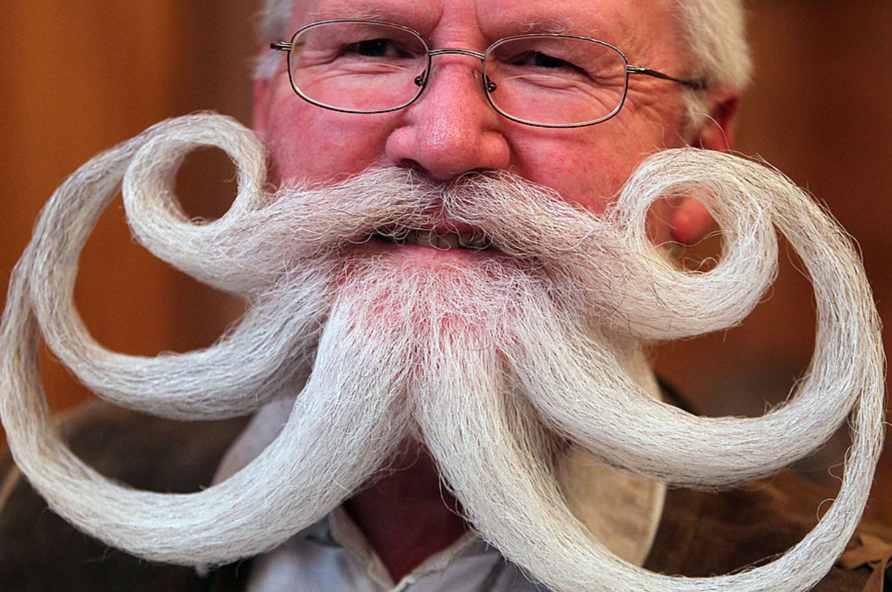 An older man with a large, elaborately styled mustache featuring multiple curl patterns smiles for the camera