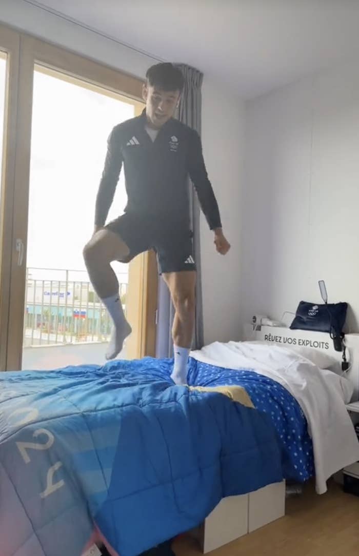 Person in sportswear and socks is jumping on a bed in a room with large windows. The bed has a blue-themed duvet cover with "RÊVEZ VOS EXPLOITS" on the headboard