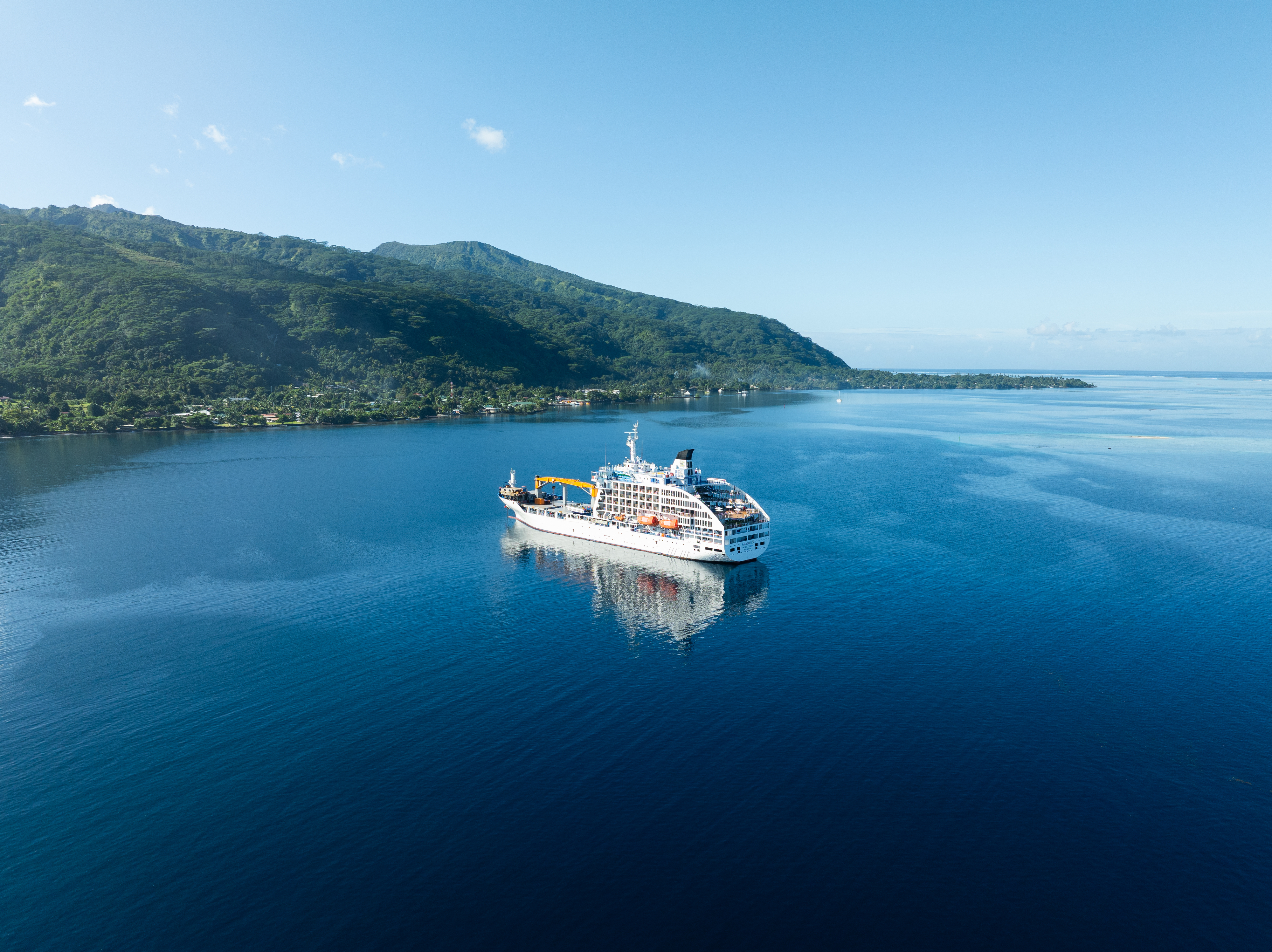 A cruise ship sails on calm blue water near a lush green island with mountains in the background, under a clear sky