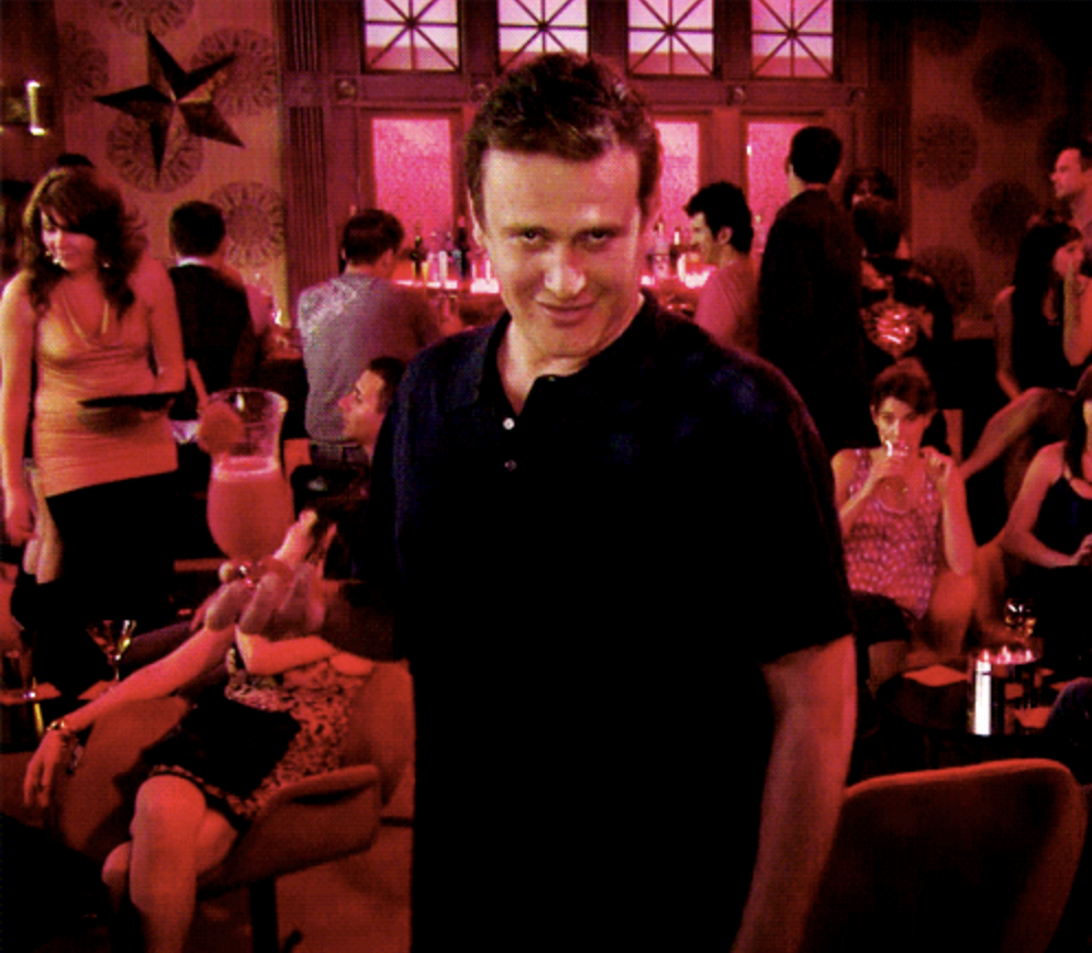 Jason Segel holding a cocktail, giving a knowing look in a lively bar setting with people mingling and enjoying drinks in the background