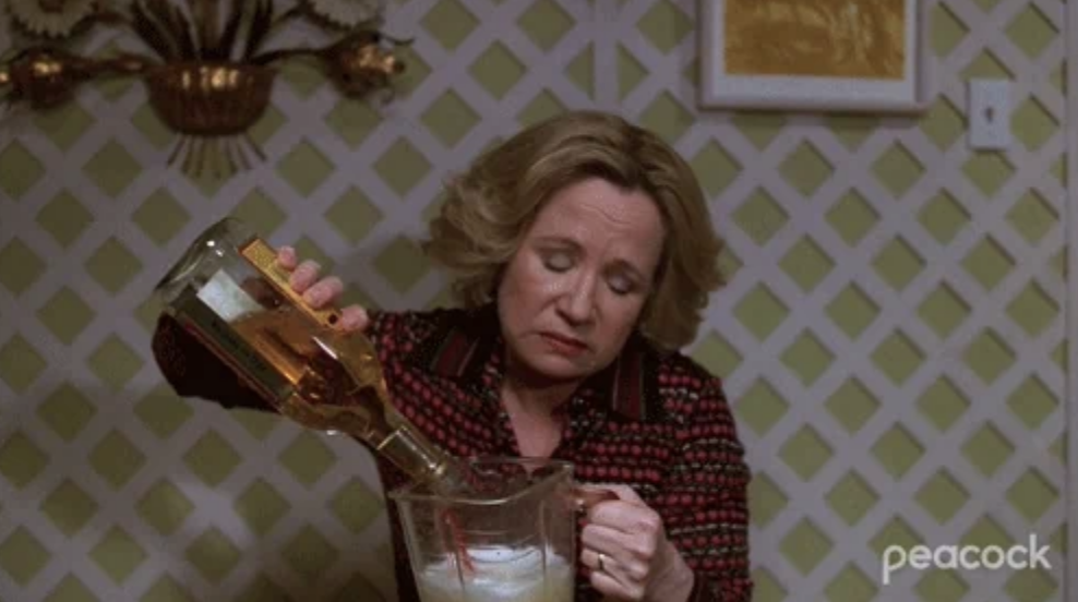 A woman is pouring alcohol into a blender in a kitchen with a green patterned wall in the background. The Peacock TV brand is visible in the corner
