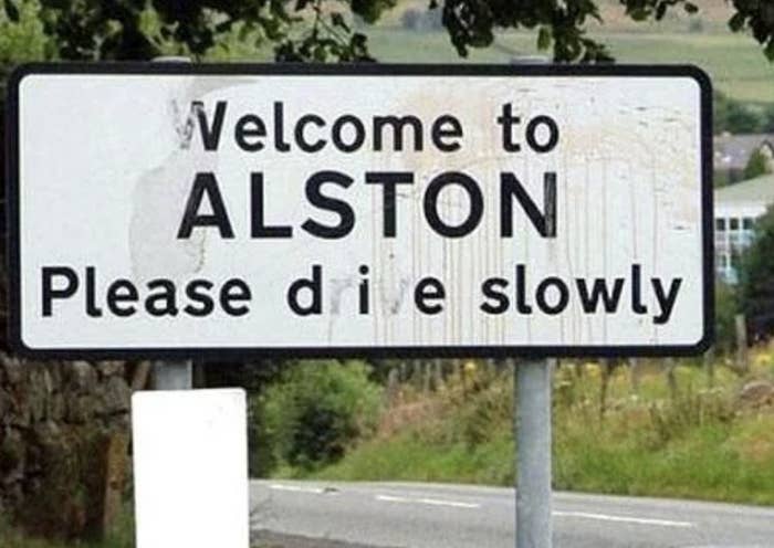 Roadside sign reads: "Welcome to ALSTON. Please drive slowly," with the letter "r" in "drive" obscured, humorously changing "drive" to "die."
