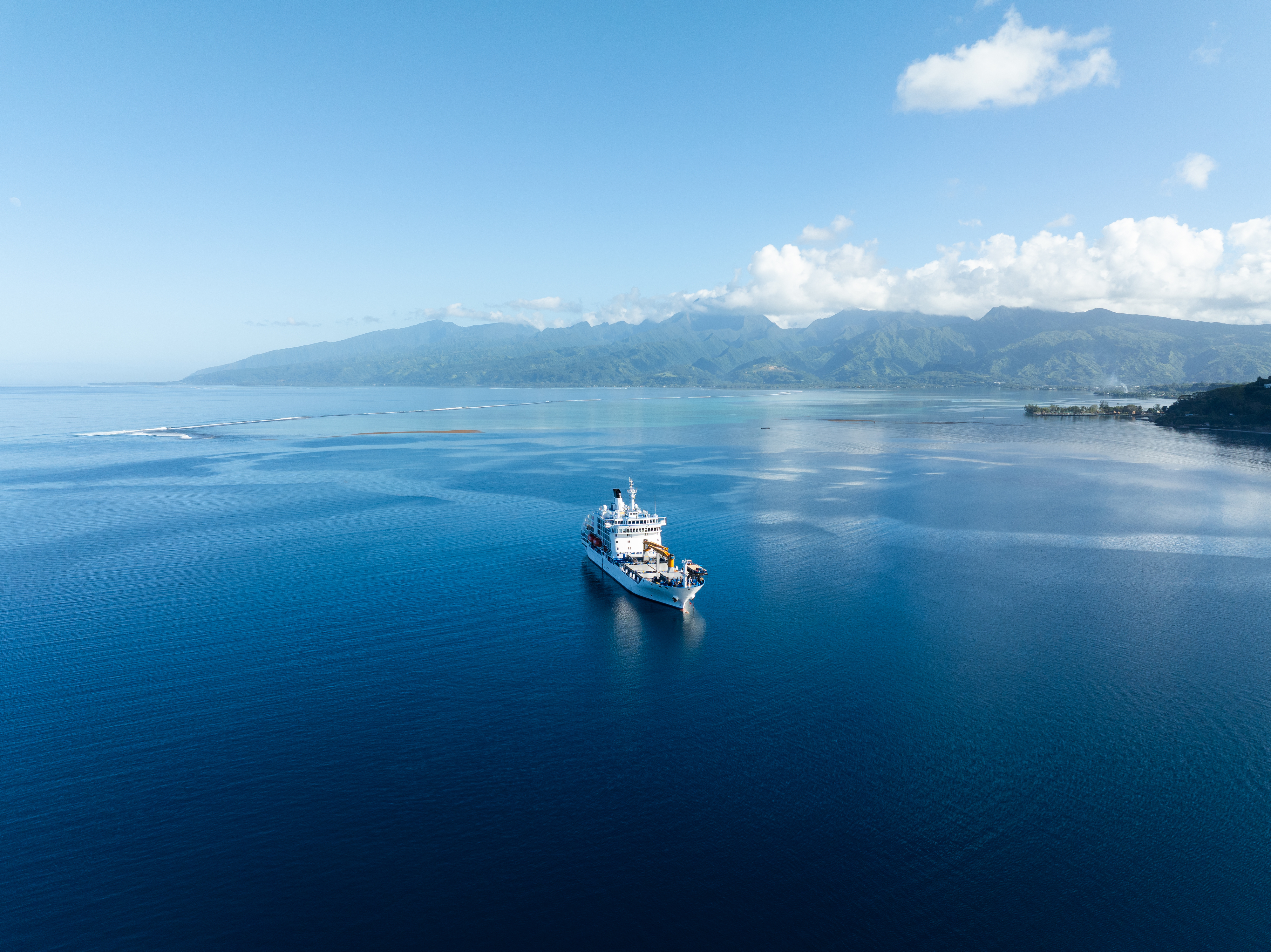 A large ship sails on calm, open water with distant mountains and a partly cloudy sky in the background