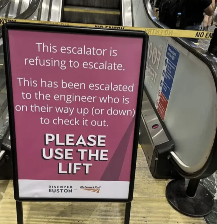 Sign near an escalator reads: "This escalator is refusing to escalate. This has been escalated to the engineer who is on their way up (or down) to check it out. PLEASE USE THE LIFT."