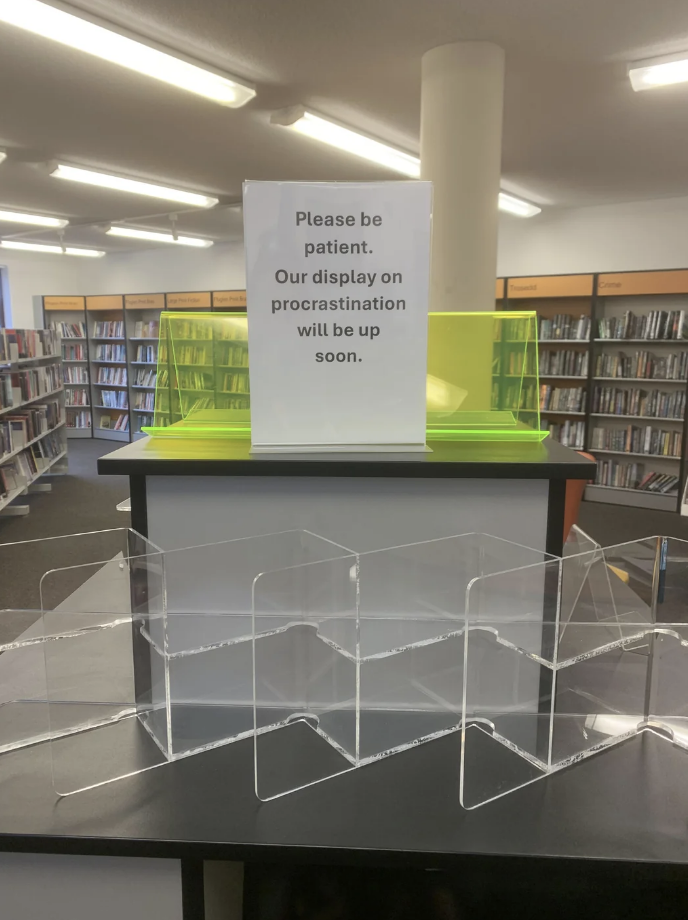 Sign in front of a library display reads, "Please be patient. Our display on procrastination will be up soon."
