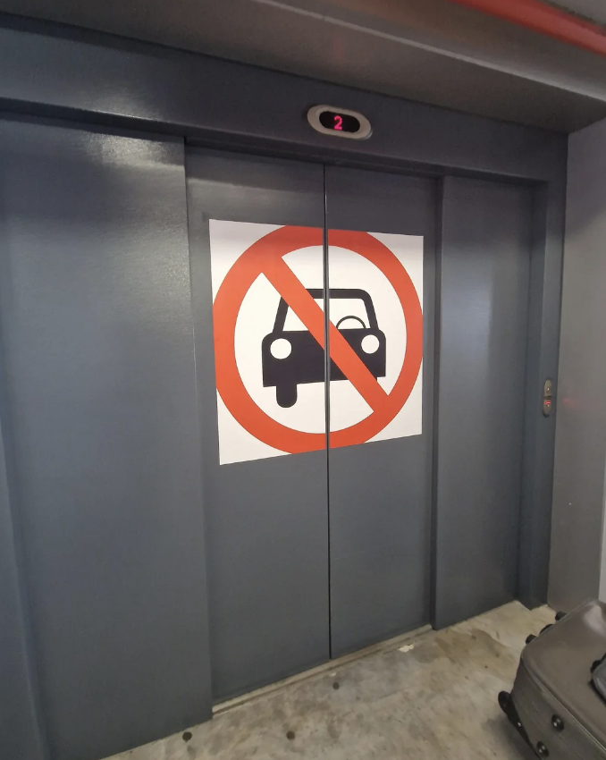 A pair of grey elevator doors with a sign showing a car symbol inside a red circle with a diagonal line through it, indicating no vehicles allowed